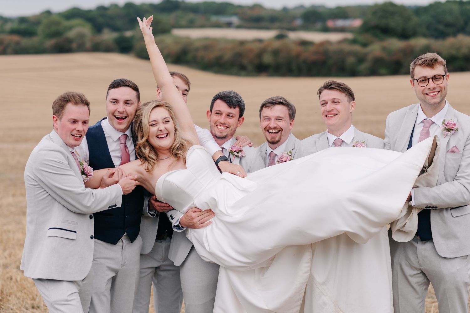 Wedding party in light gray suits lifts bride in white dress during outdoor photo session.