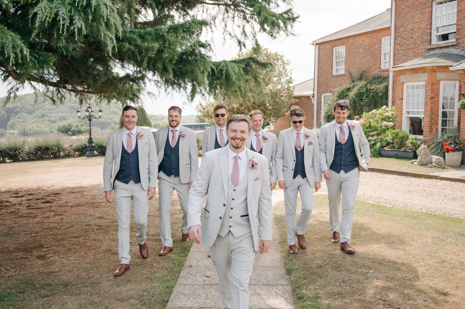 Wedding party members in light gray suits and pink accessories walking together outdoors by a brick building.