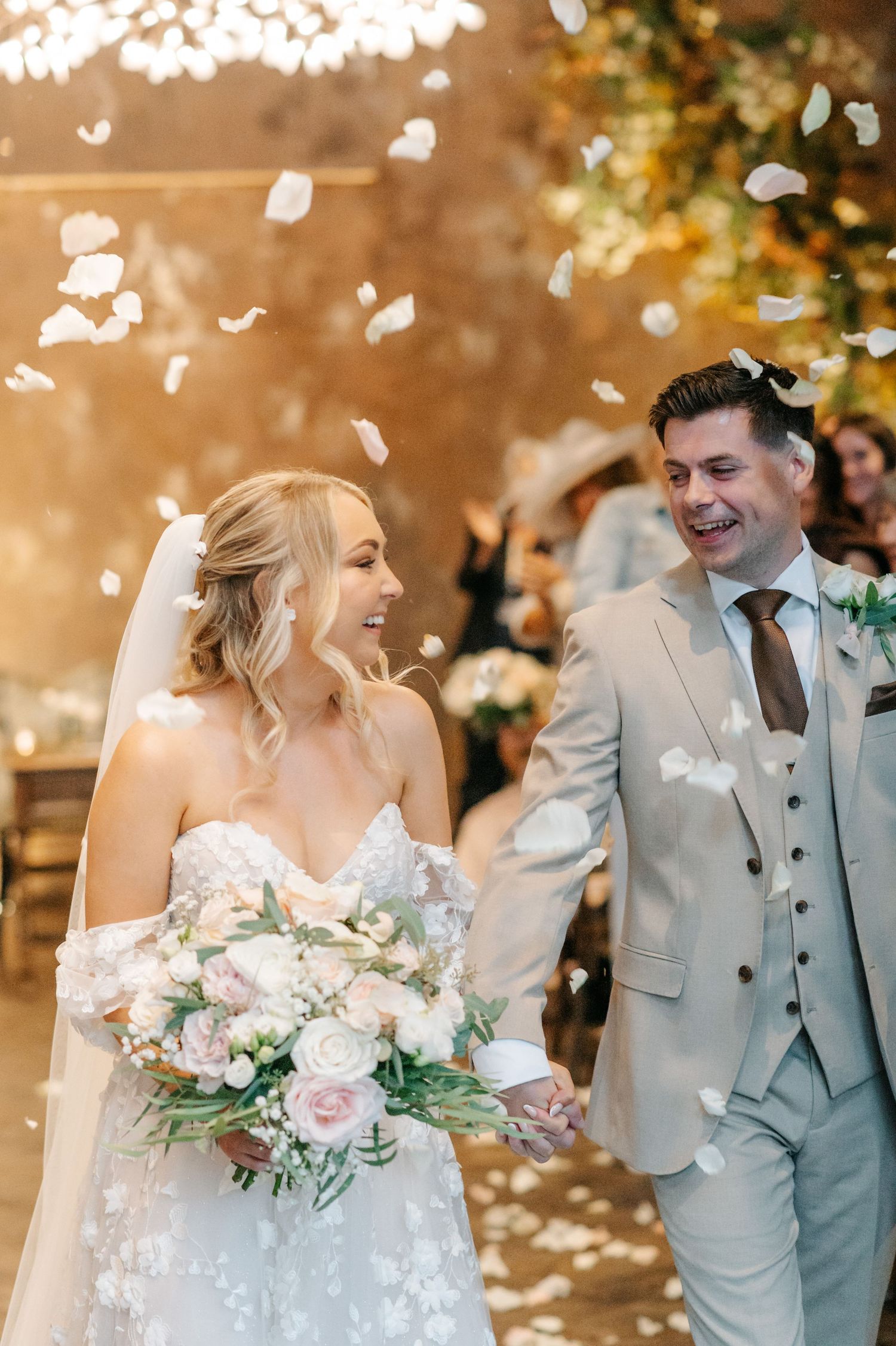 Wedding couple laughs joyfully as white flower petals fall around them during romantic outdoor celebration.