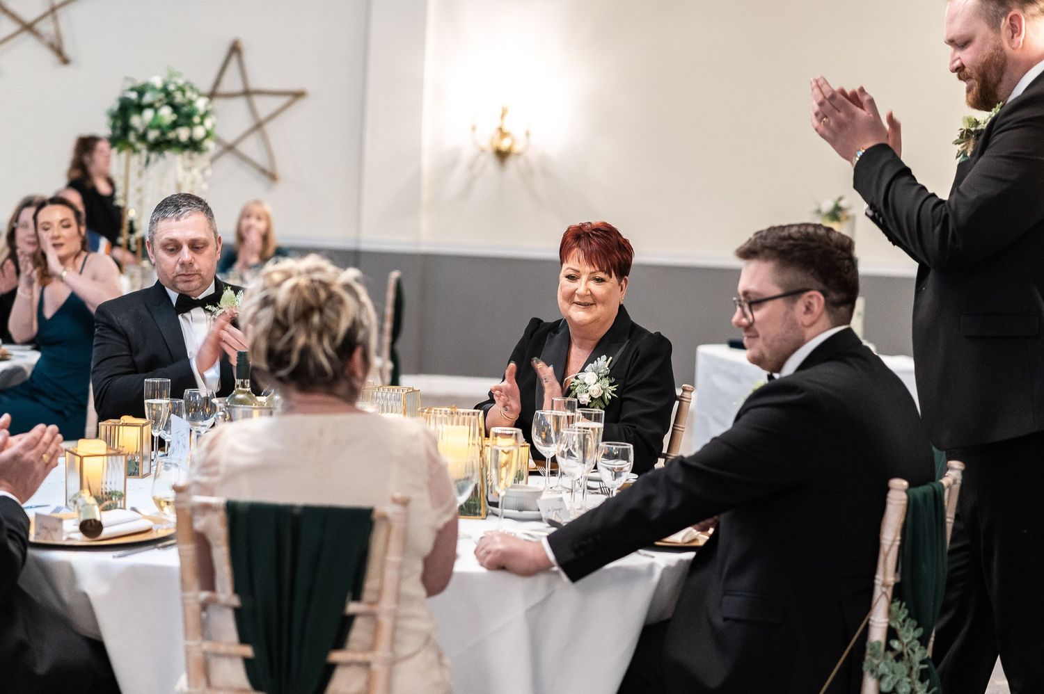 Wedding reception guests seated at elegant tables with a person standing and giving a toast.