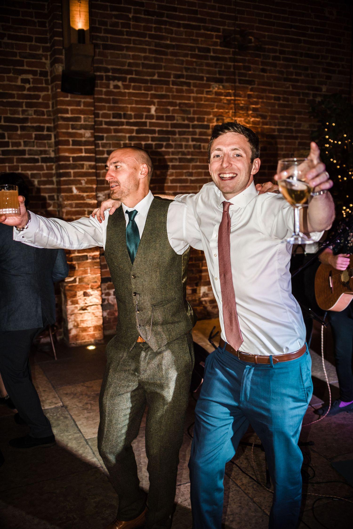 Two guests dance enthusiastically at a wedding reception in a brick-walled venue.