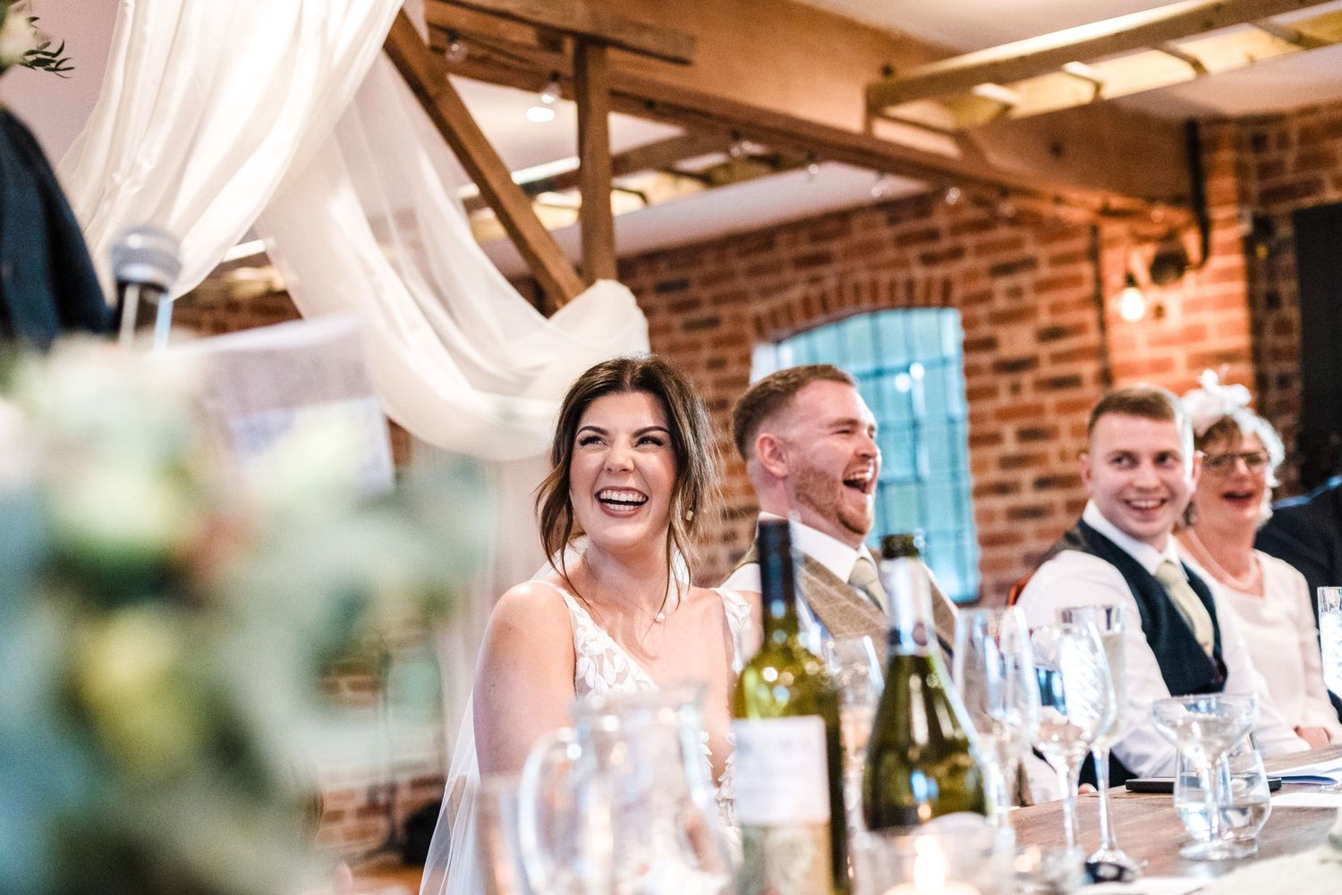 Wedding guests enjoying speeches during a reception dinner in a rustic brick venue with white draping.