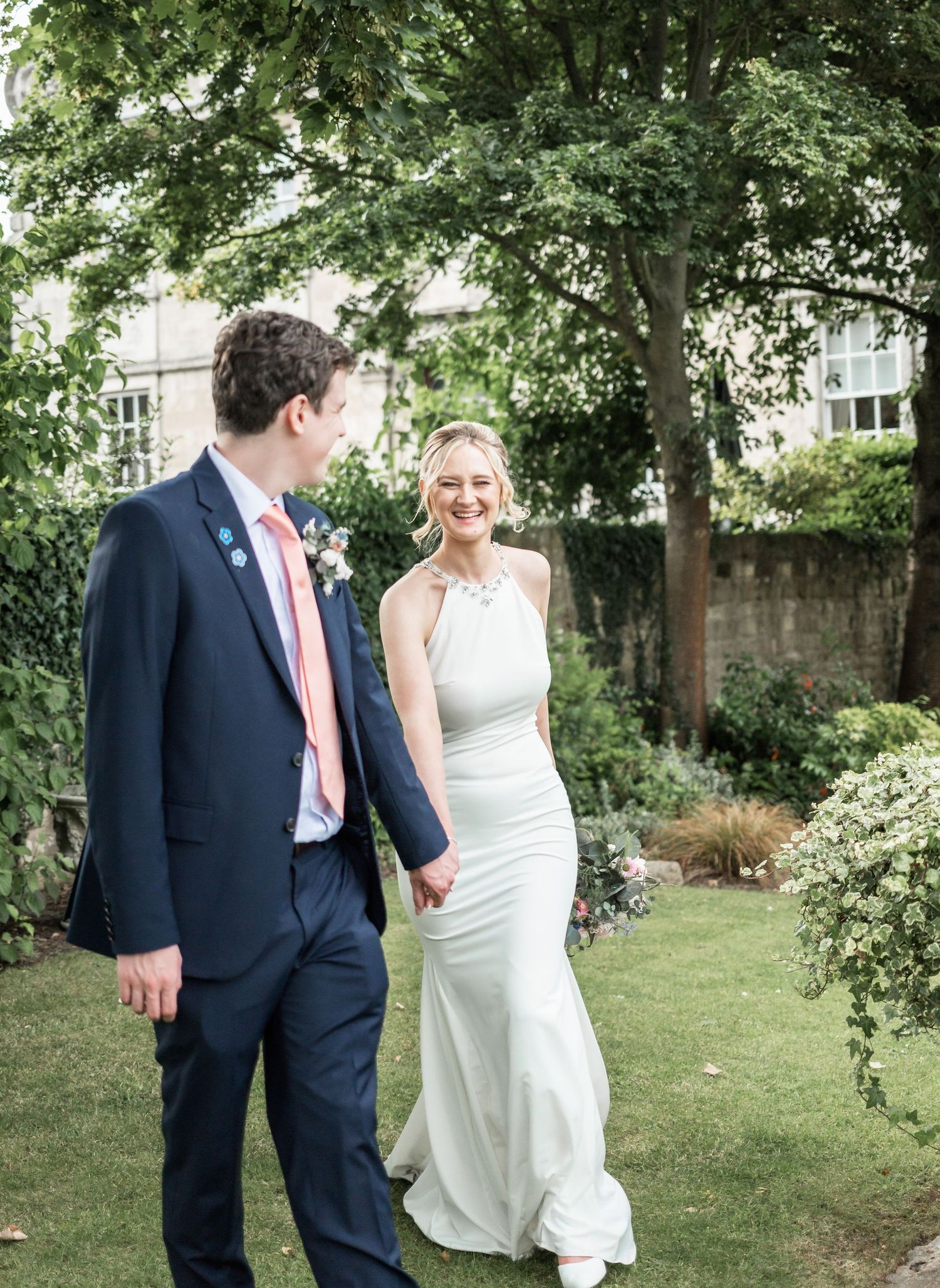 A wedding couple poses together in a garden setting with lush green trees and landscaping.