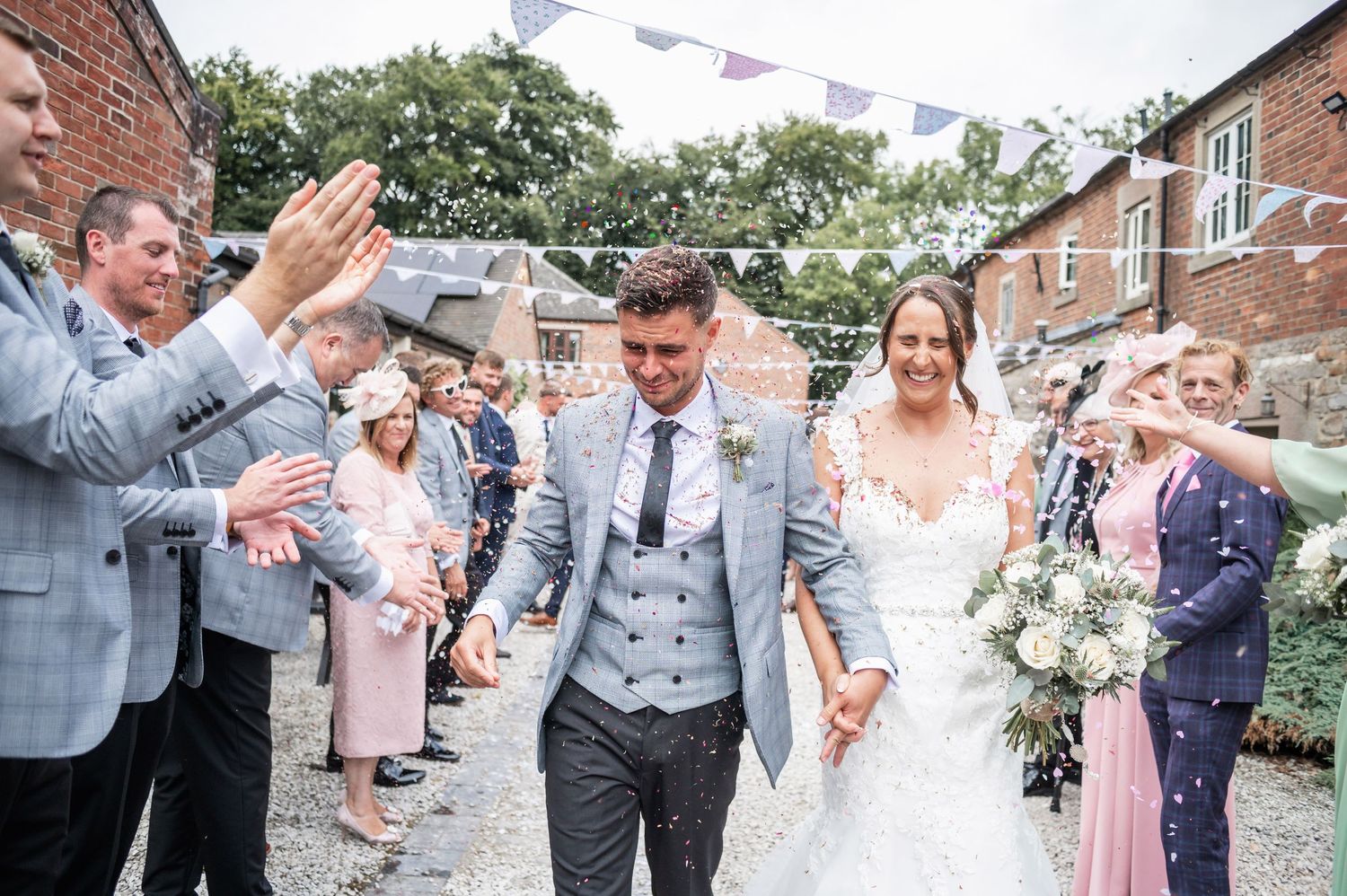 Wedding couple walks through crowd of clapping guests on cobblestone path.