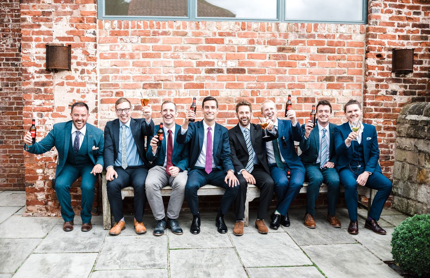 Groomsmen in blue suits pose together on stone bench against brick wall.