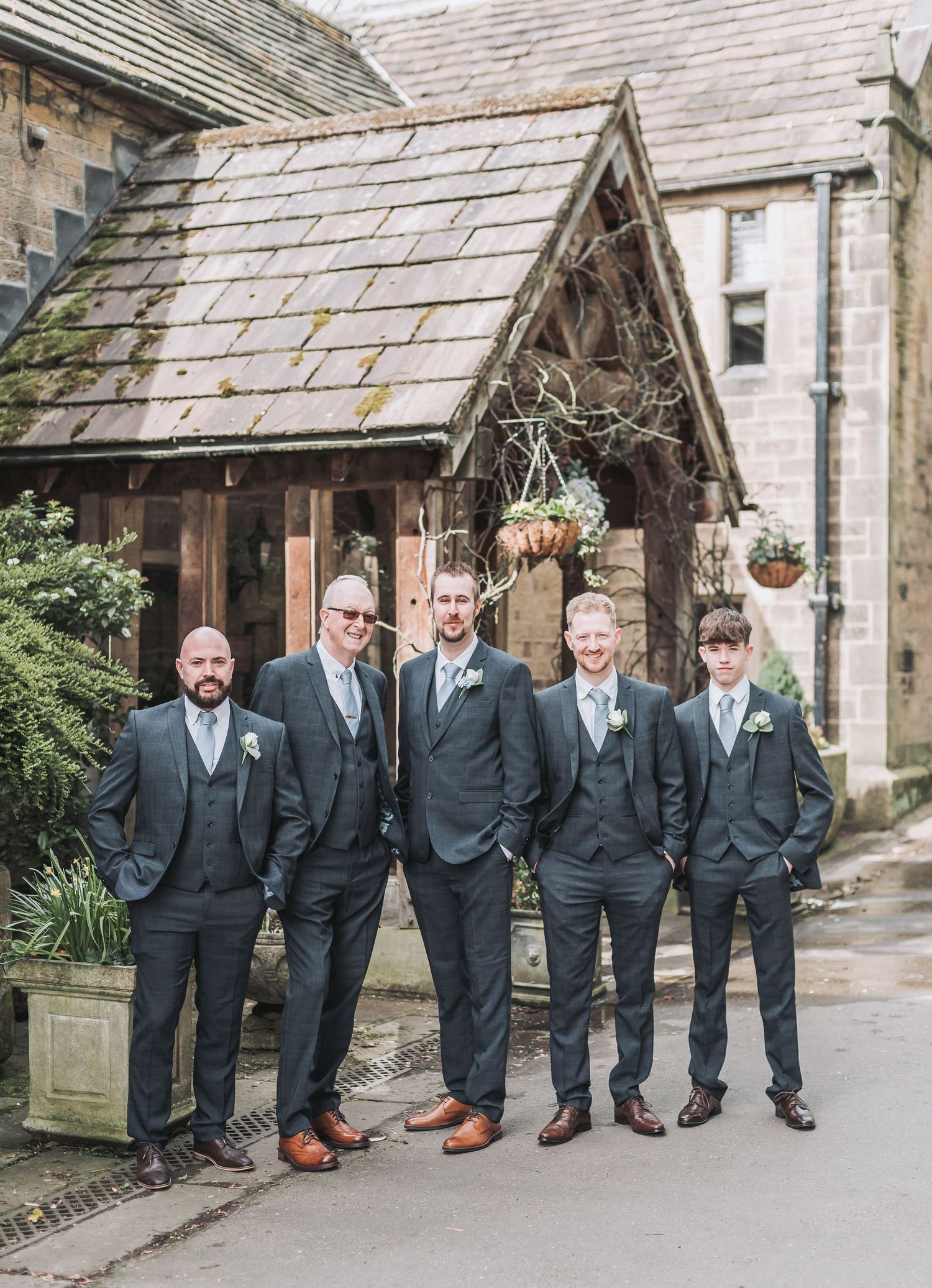Five groomsmen in dark suits and brown shoes pose outside a stone church entrance.