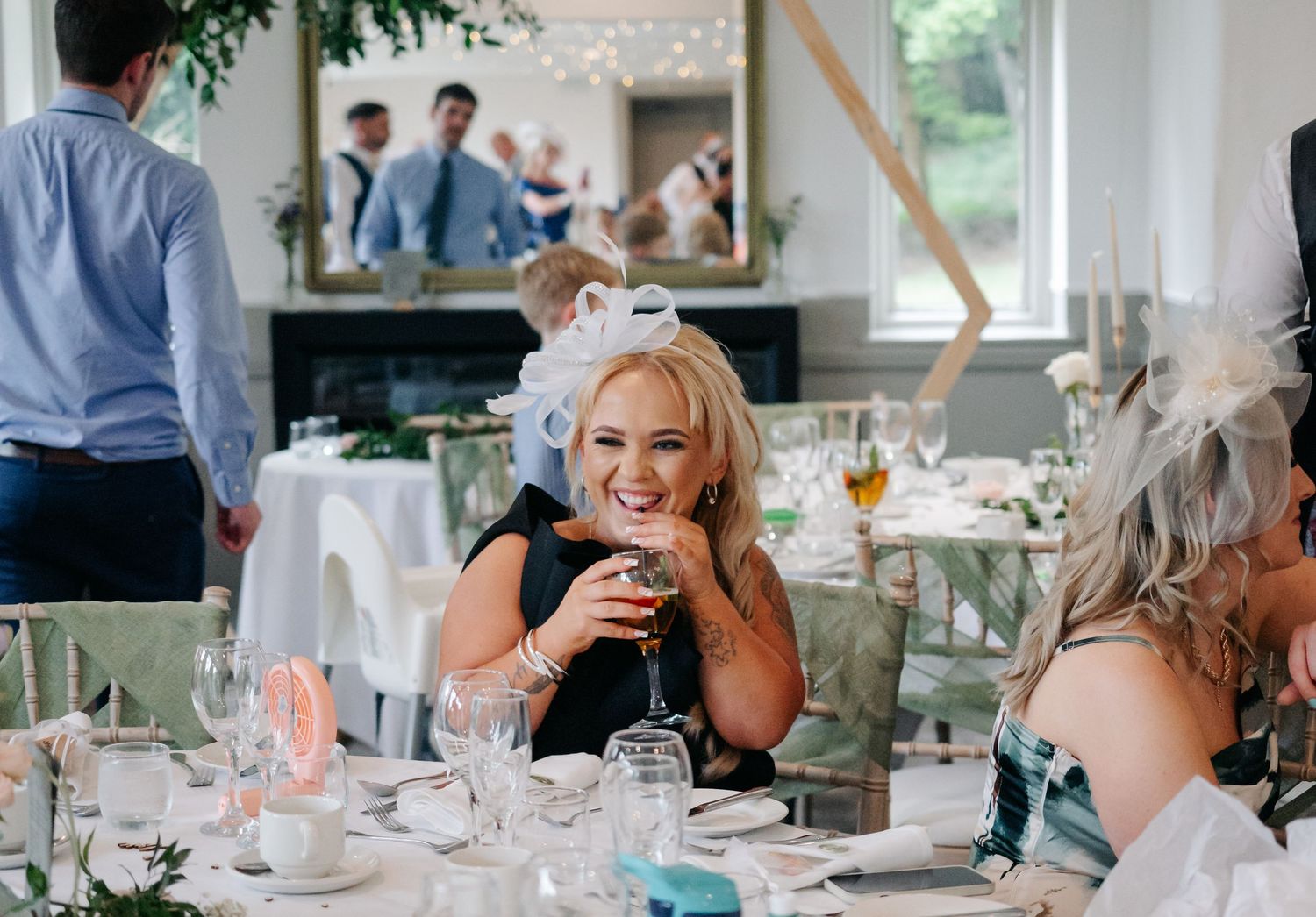 Wedding guest in black dress laughs while holding drink at reception dinner table with decorative place settings.