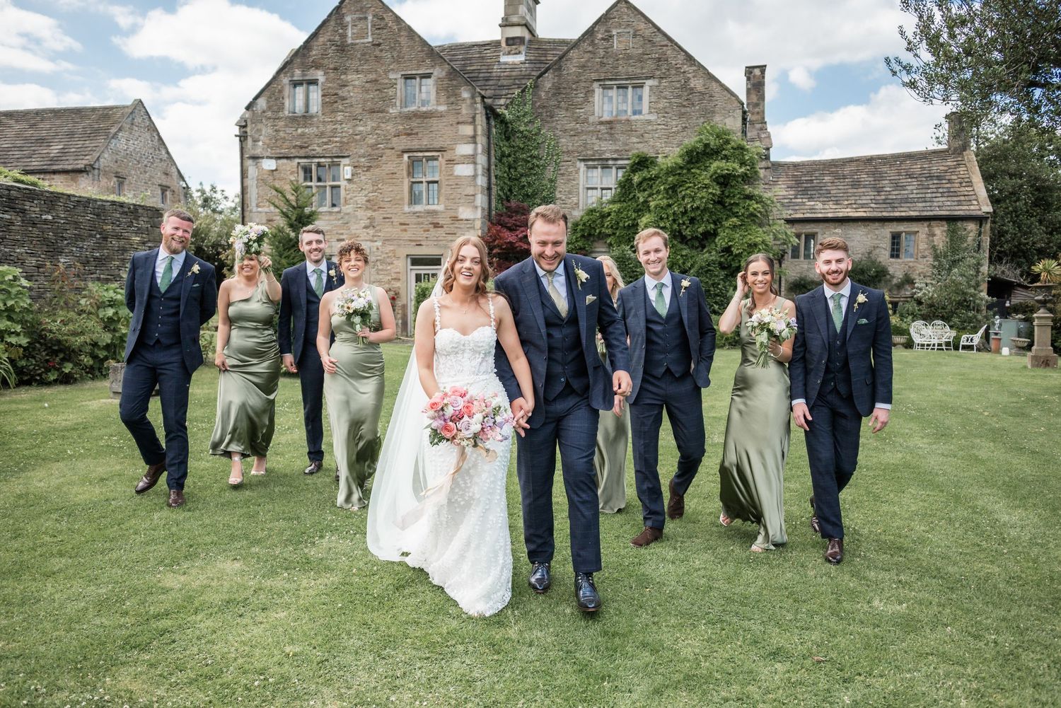 Wedding party walks and laughs together across a lush garden lawn with stone manor house in background.