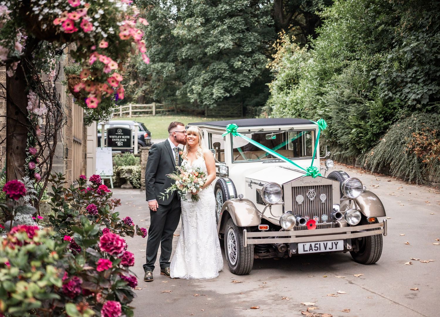Newlyweds pose with classic vintage wedding car surrounded by pink flowers.