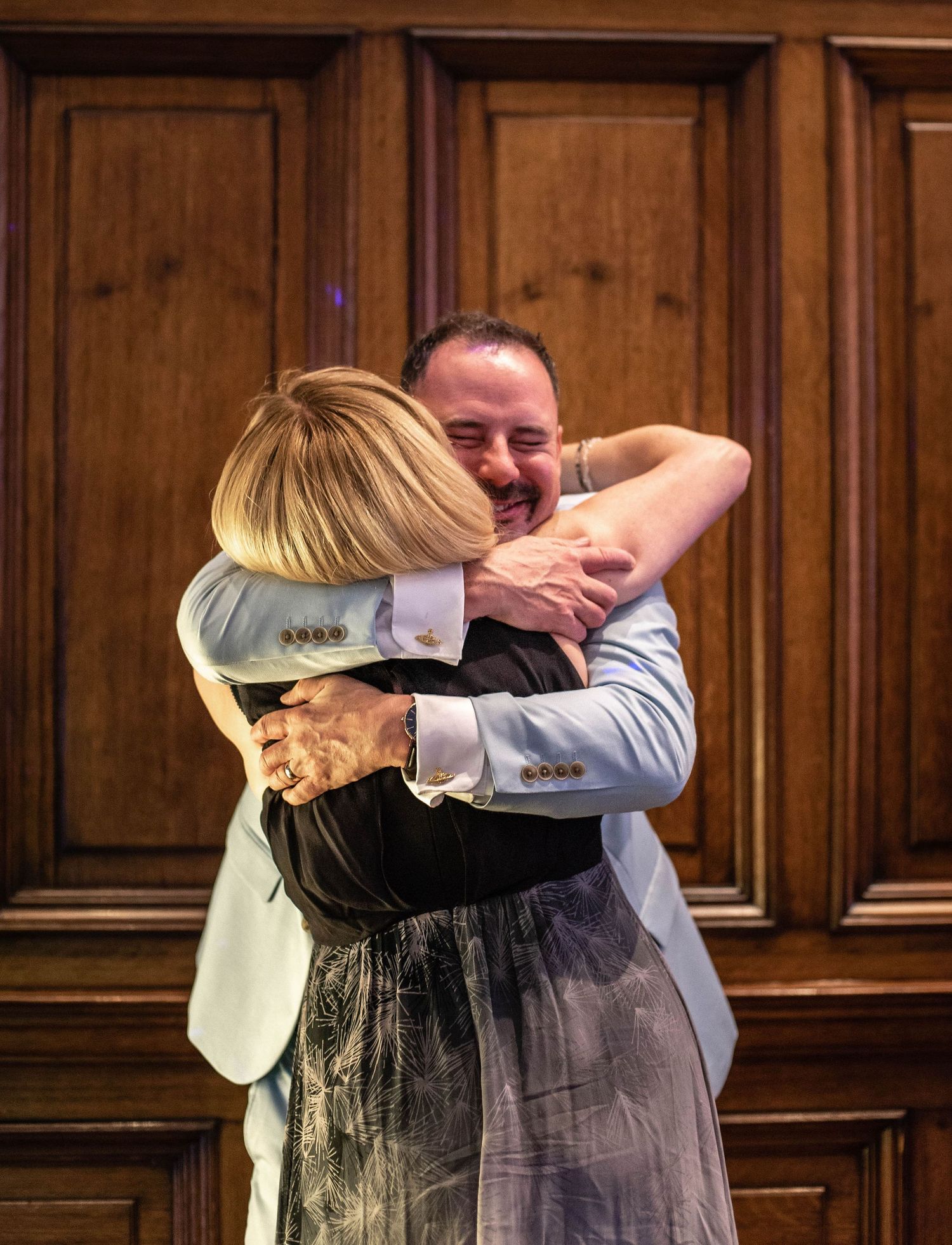 An emotional embrace between two people at a formal event against wooden paneled walls.