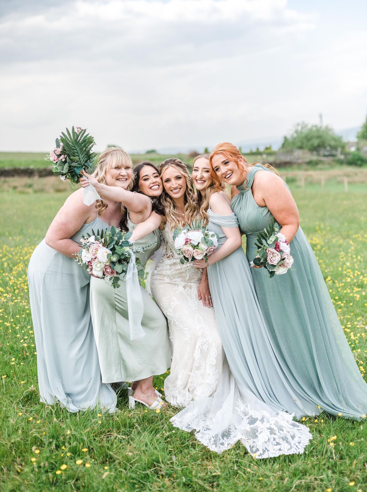 Bridesmaids in sage green dresses pose playfully with the bride in a flower-filled meadow.