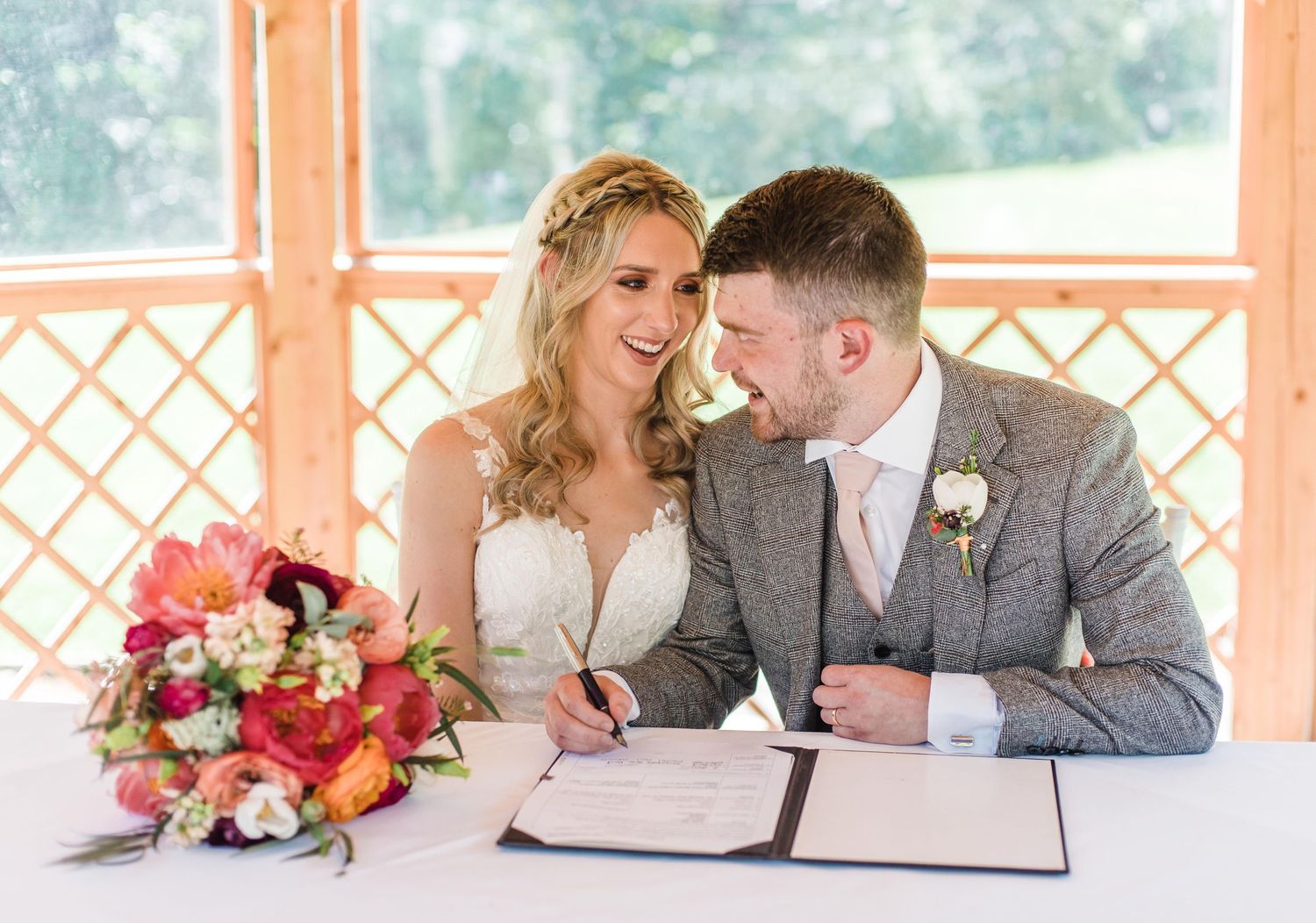 Couple signs marriage documents at wooden table with colorful floral arrangement.