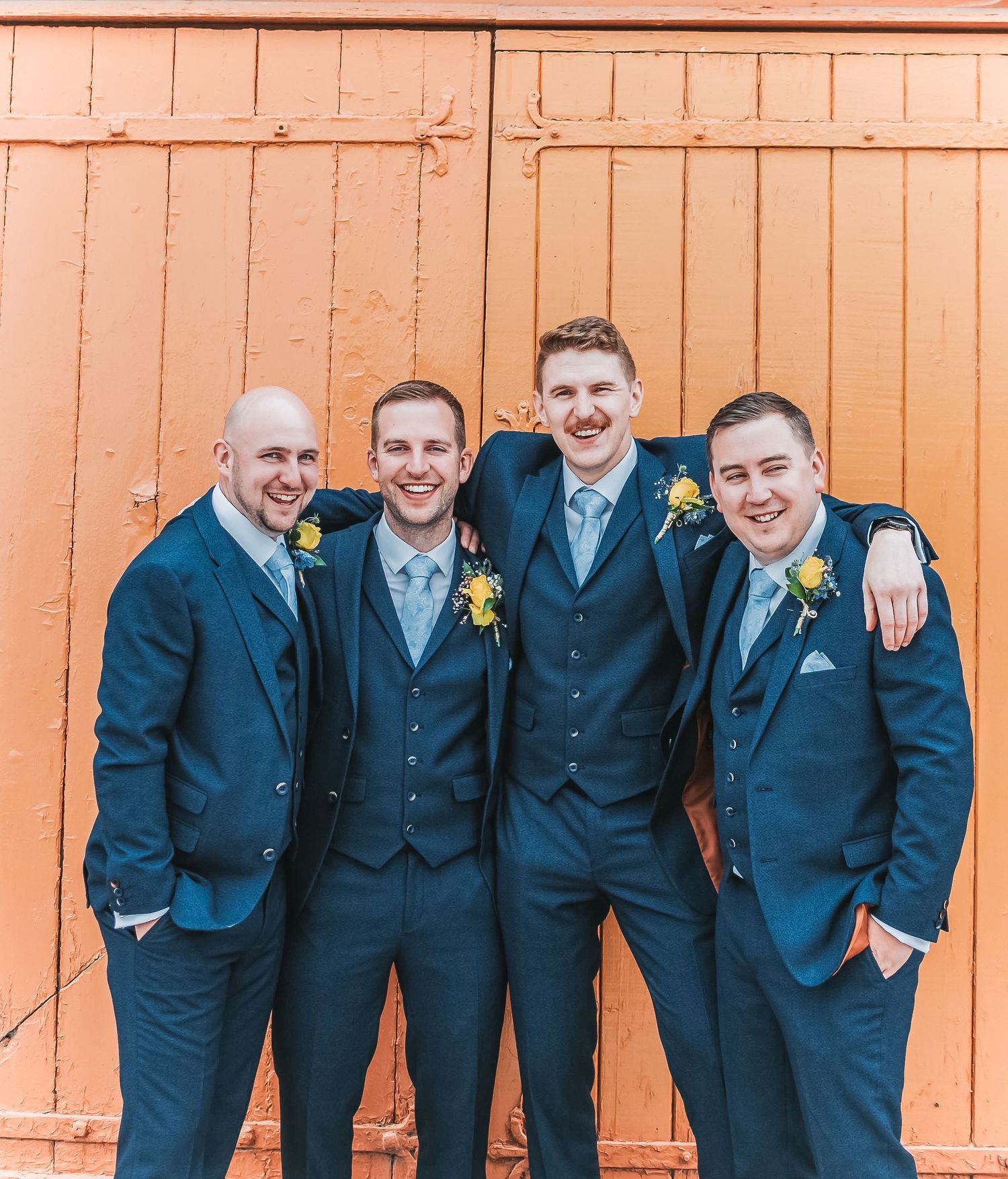 Groomsmen in navy suits posing against orange wooden door at wedding venue.