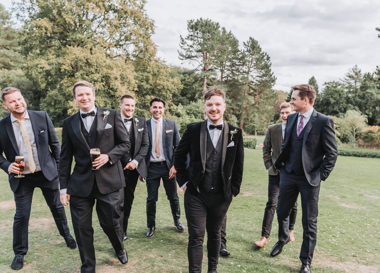 Groomsmen in black suits walk across a sunny lawn while sharing laughs during an outdoor wedding celebration.