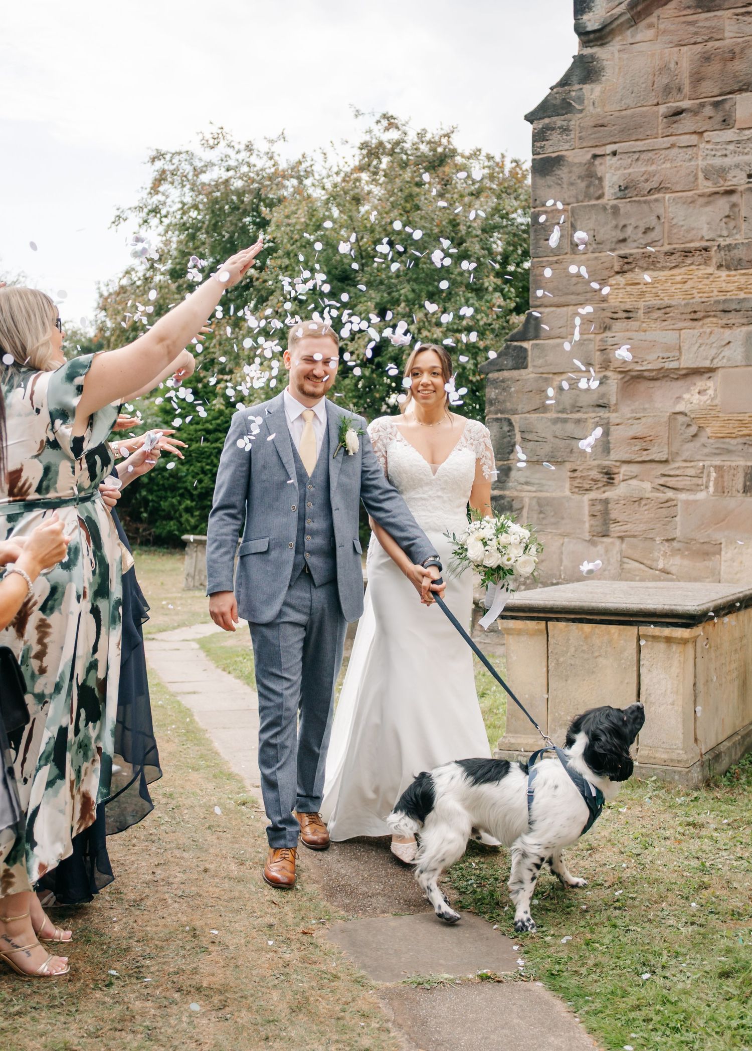 A couple and their dog exit the church while guests throw confetti.