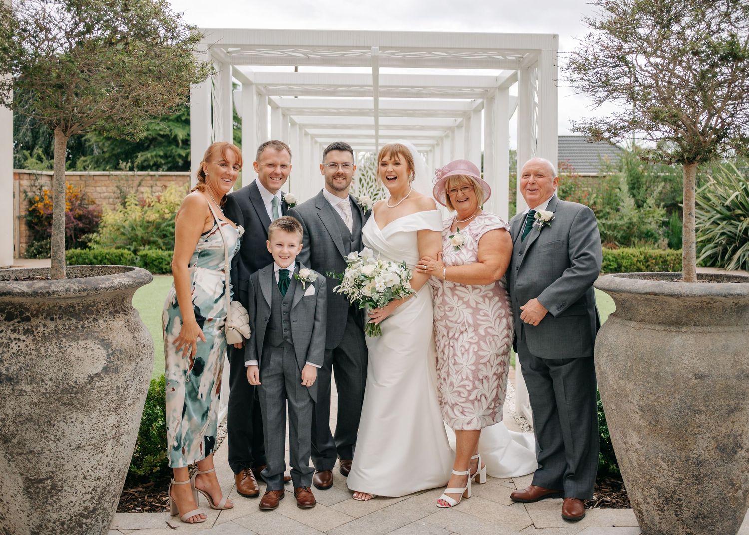 Wedding party members gathered under a white pergola for formal photographs.