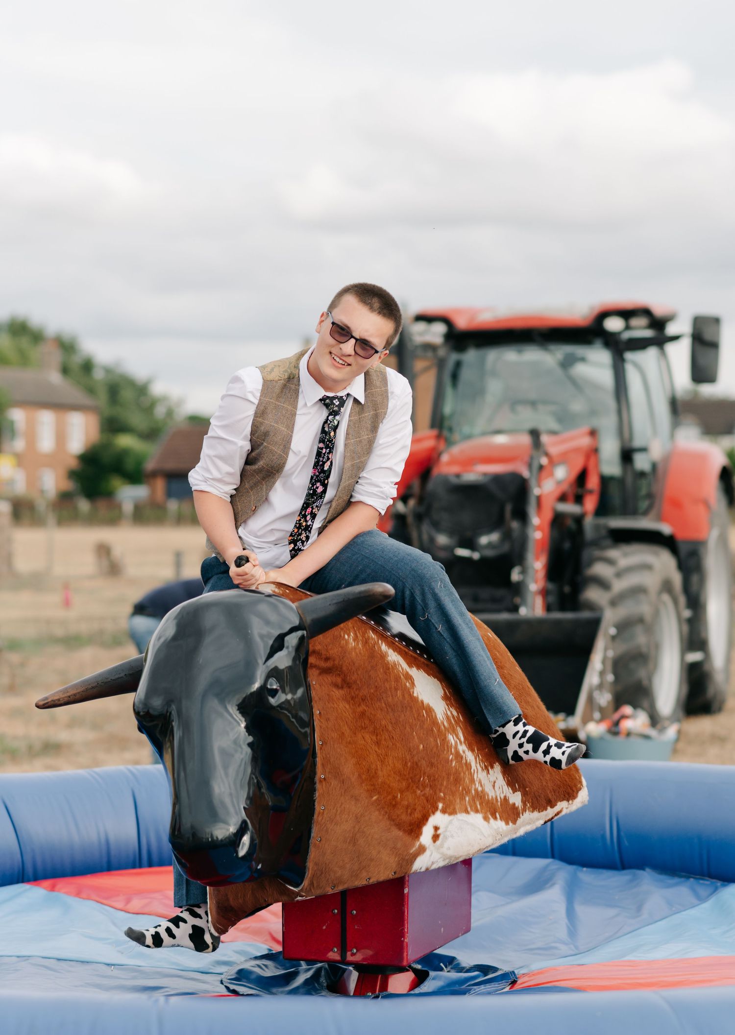 Guest rides mechanical bull at outdoor country wedding reception with tractors in background.