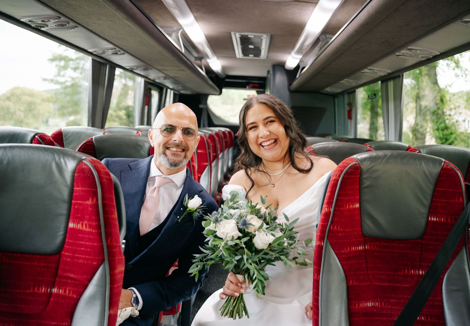 A happy couple sits together on red seats inside a luxury tour bus on their wedding day.