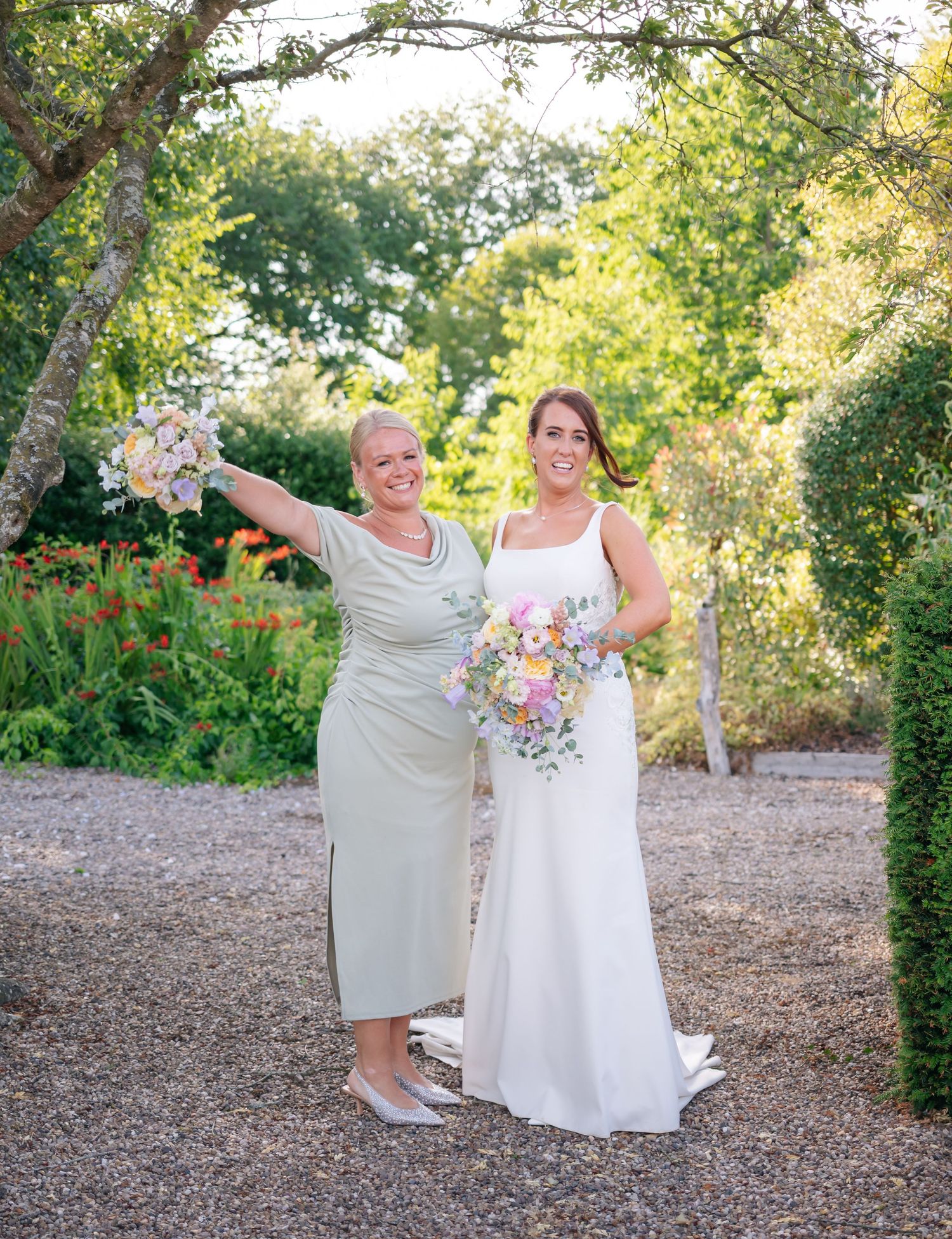 Two people in light colored attire pose playfully on a gravel path surrounded by greenery.