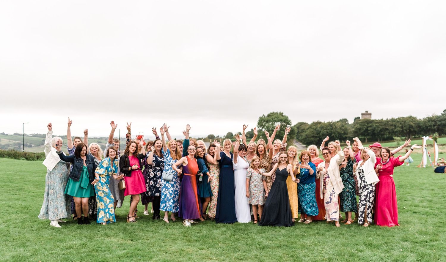 Large group of wedding guests in colorful dresses and formal attire posing together on a green lawn.