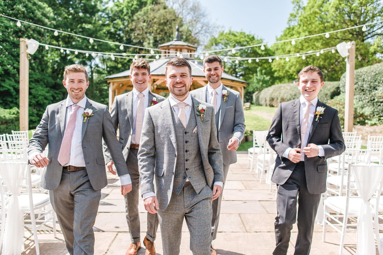 Groomsmen in matching grey suits walk down outdoor ceremony aisle.