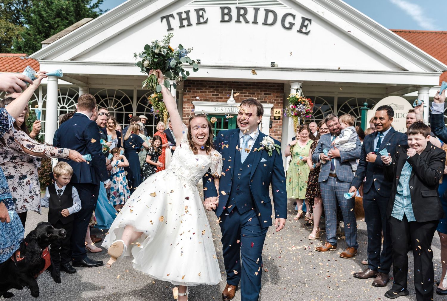 Newlyweds celebrate outside The Bridge venue as wedding guests throw confetti.