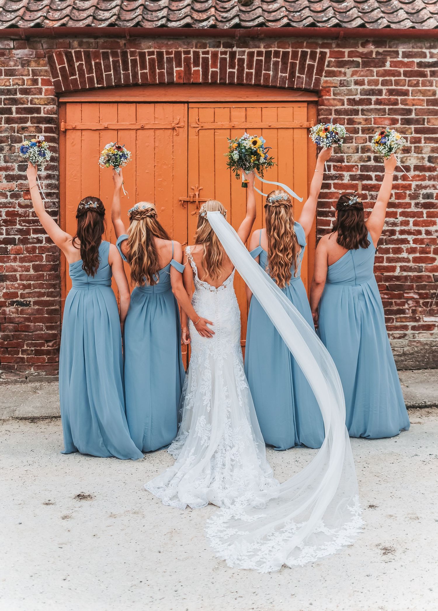 Wedding party posed with backs turned showing dusty blue bridesmaid dresses against orange barn door with floral decor.