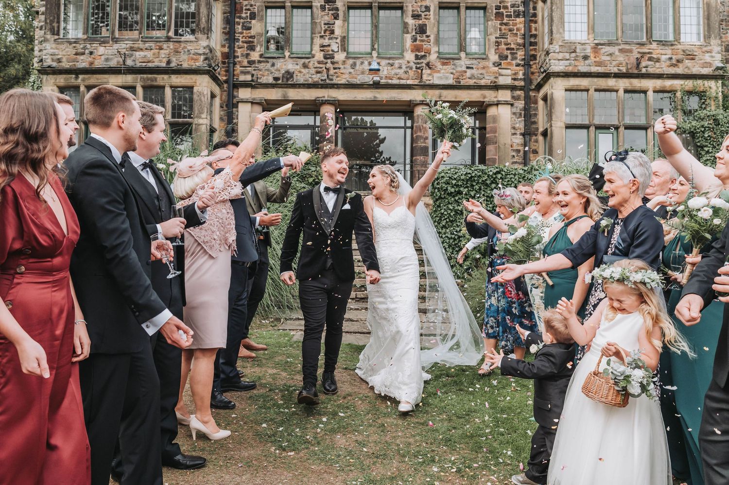 Wedding guests throw confetti as couple walks through on lawn of historic building.