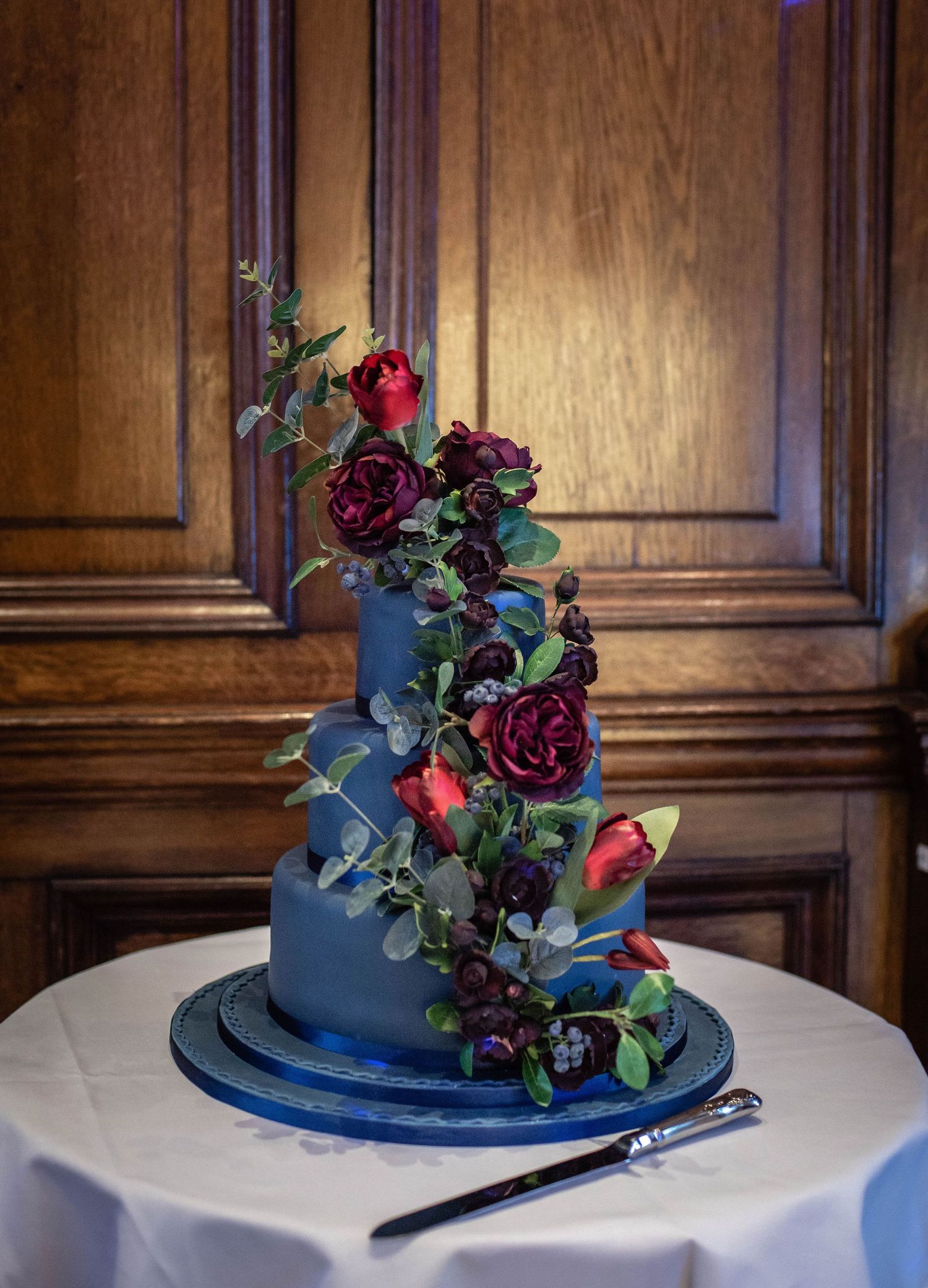 A navy blue wedding cake decorated with cascading dark red roses sits on a white tablecloth.