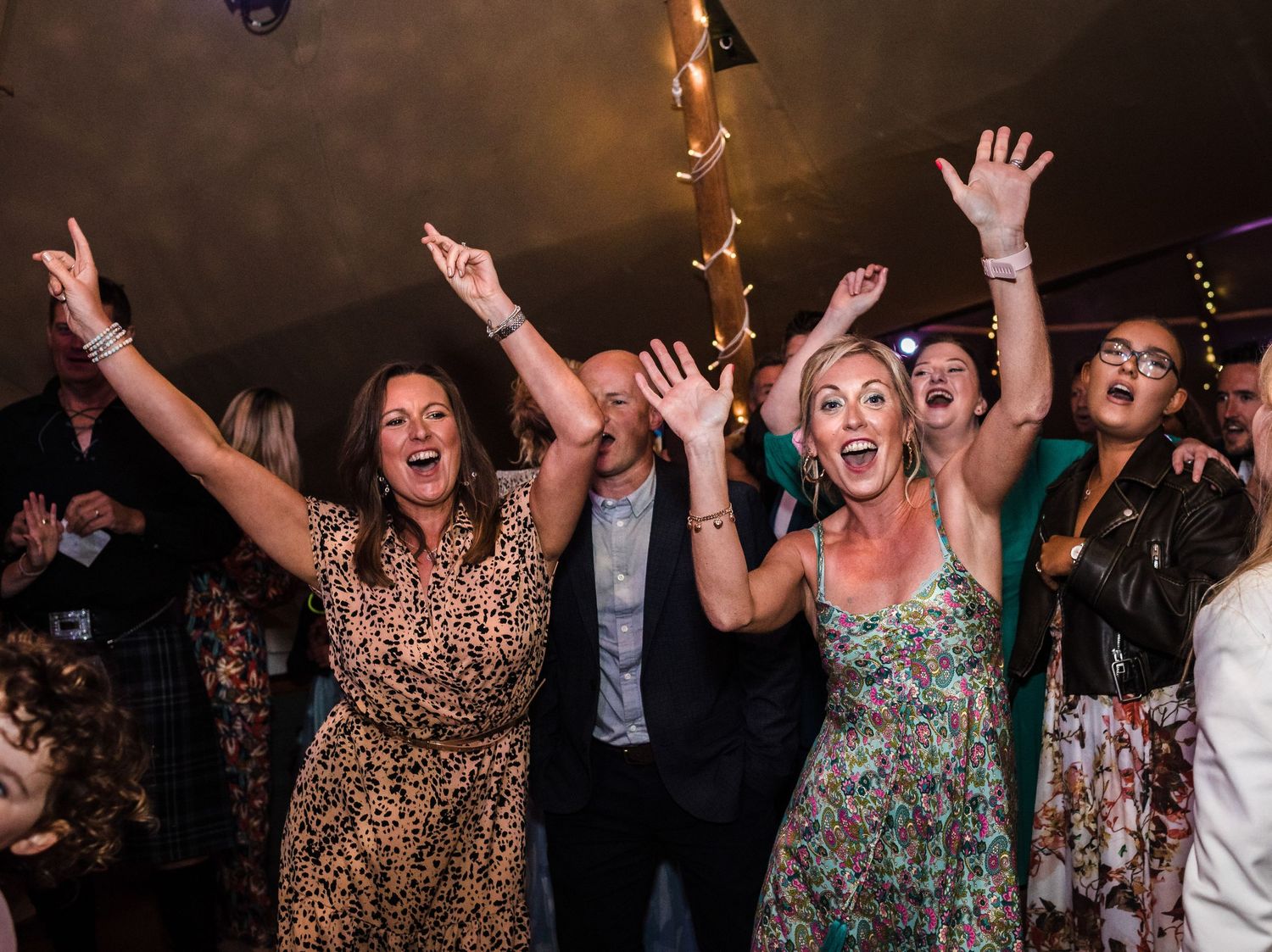 Crowd of wedding guests dancing enthusiastically with raised arms under string lights at an indoor reception.