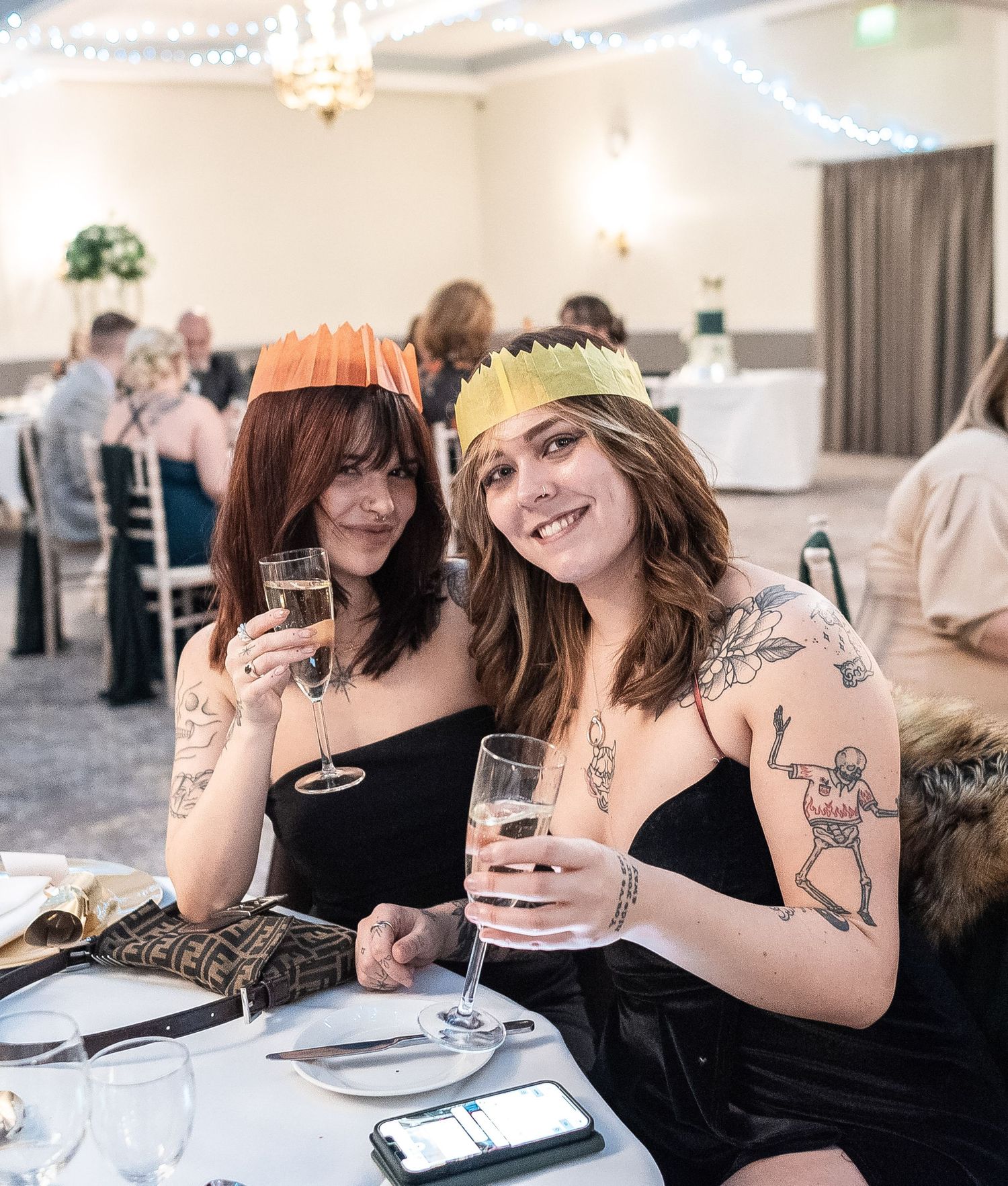 Wedding guests in black dresses wearing party crowns toast with champagne at reception.