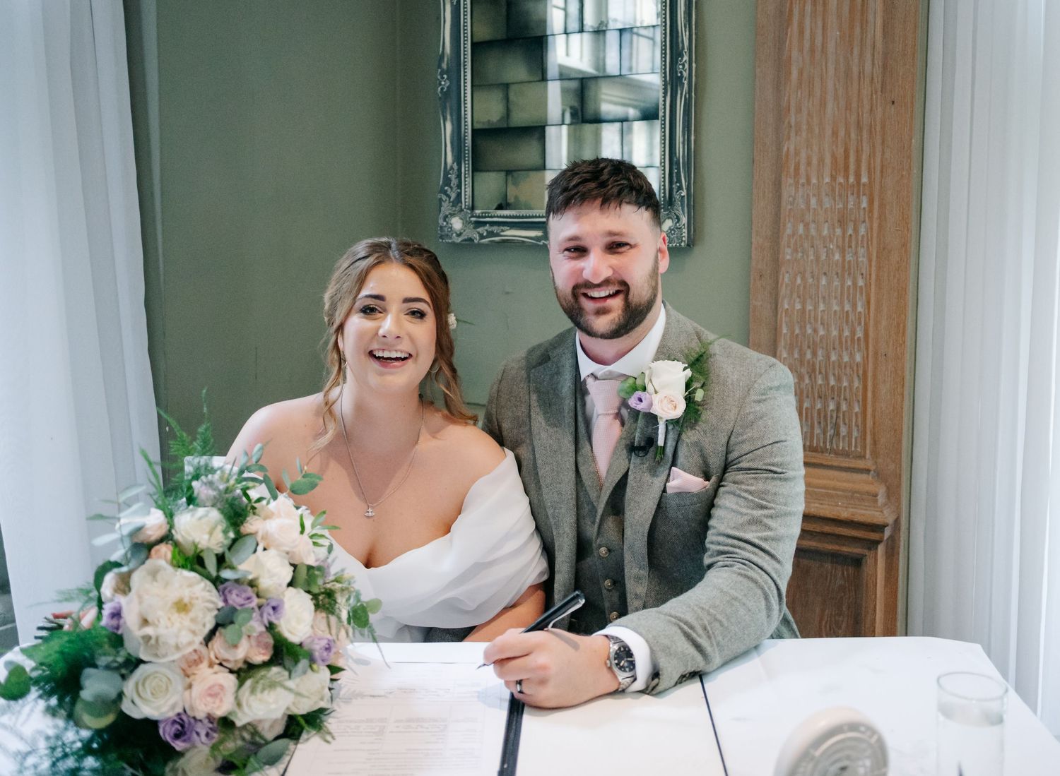 A couple sits together at a table during their wedding, with a soft pink and white floral arrangement in the foreground.