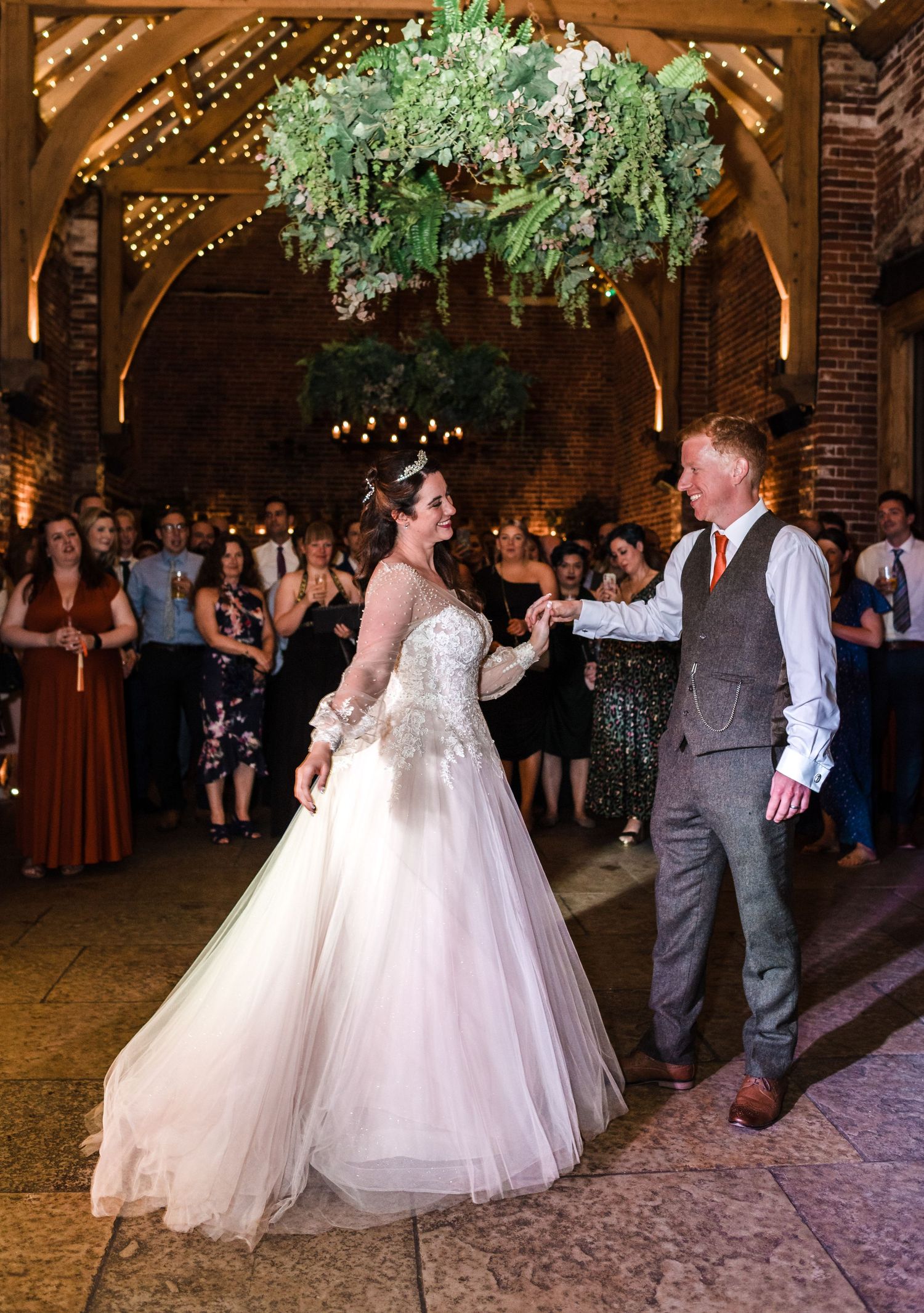 A wedding couple dances under a lush greenery chandelier in a beautifully lit brick venue.