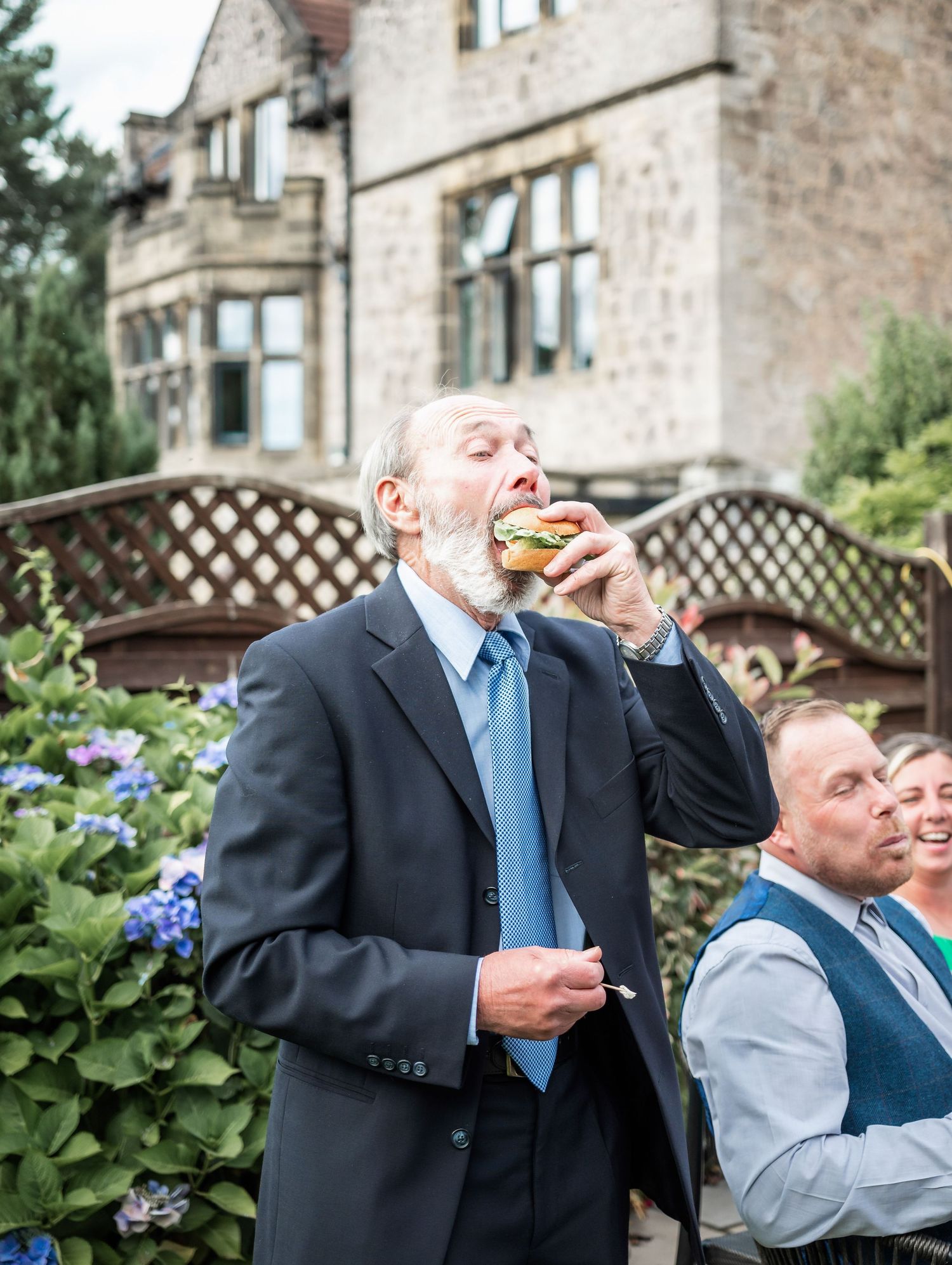 Guest in navy suit enjoying a burger at an outdoor garden reception.