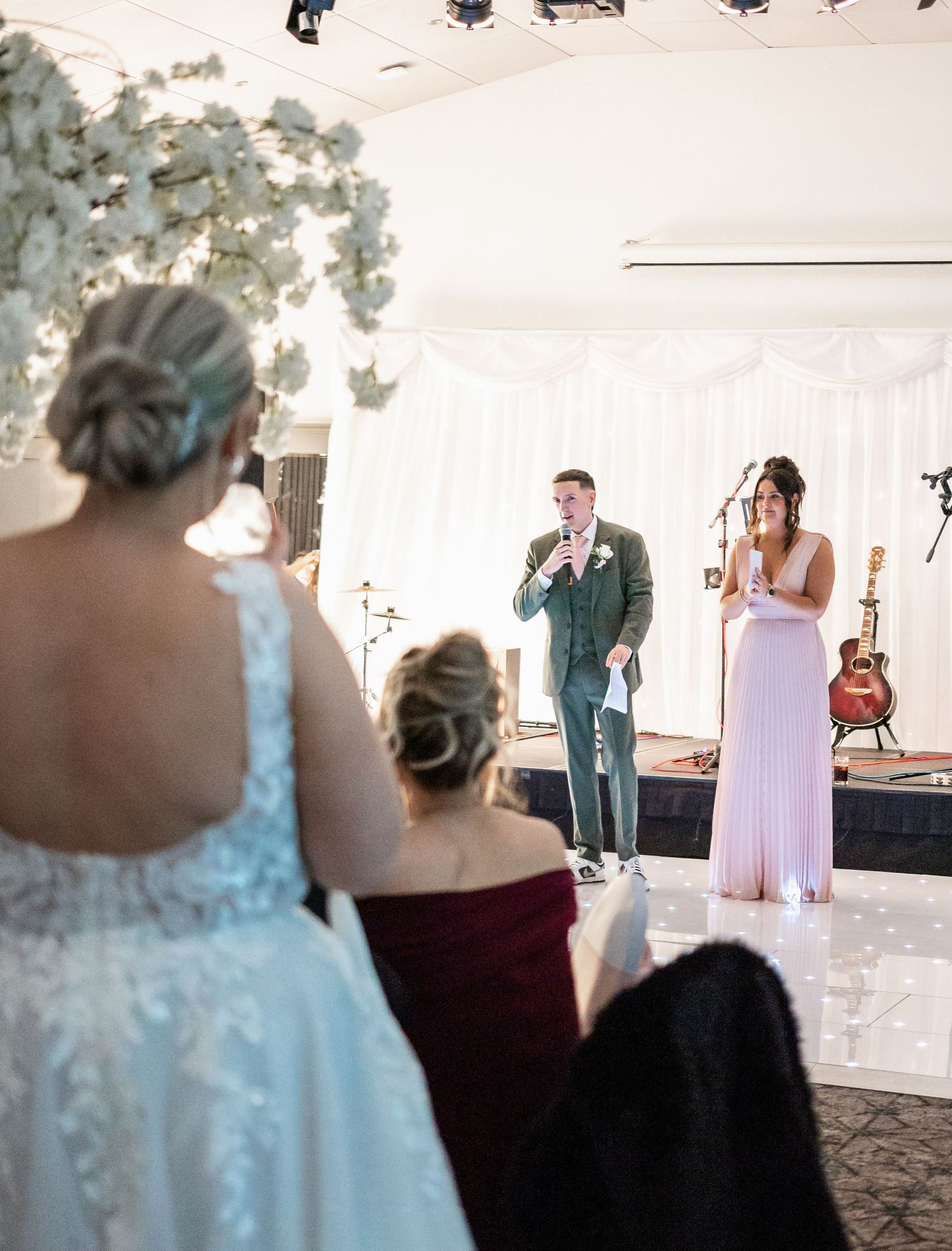 Wedding party members give speeches on stage decorated with white floral arrangements.