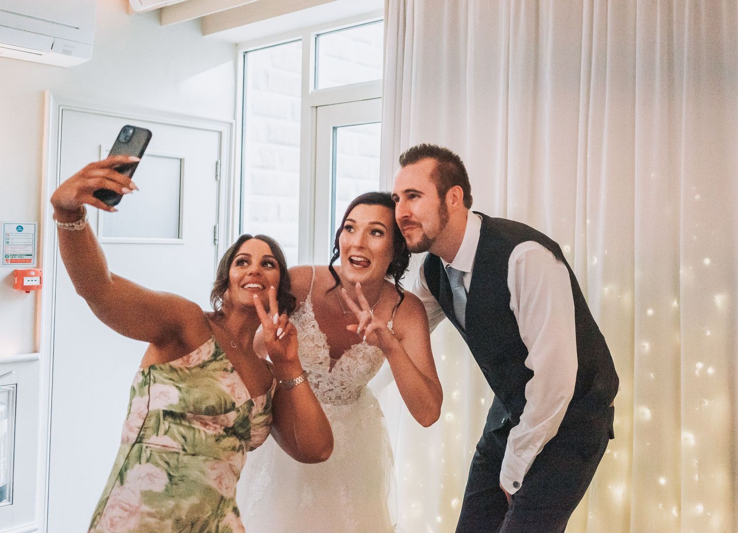Wedding party taking a celebratory selfie together in bright indoor venue with string lights.