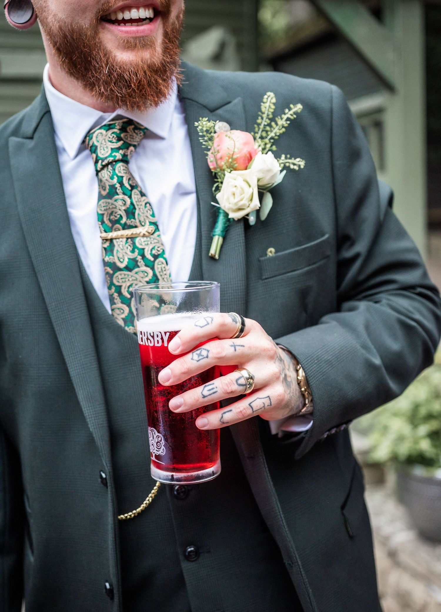 Groom in green suit with floral tie and boutonniere holds red drink at outdoor wedding.