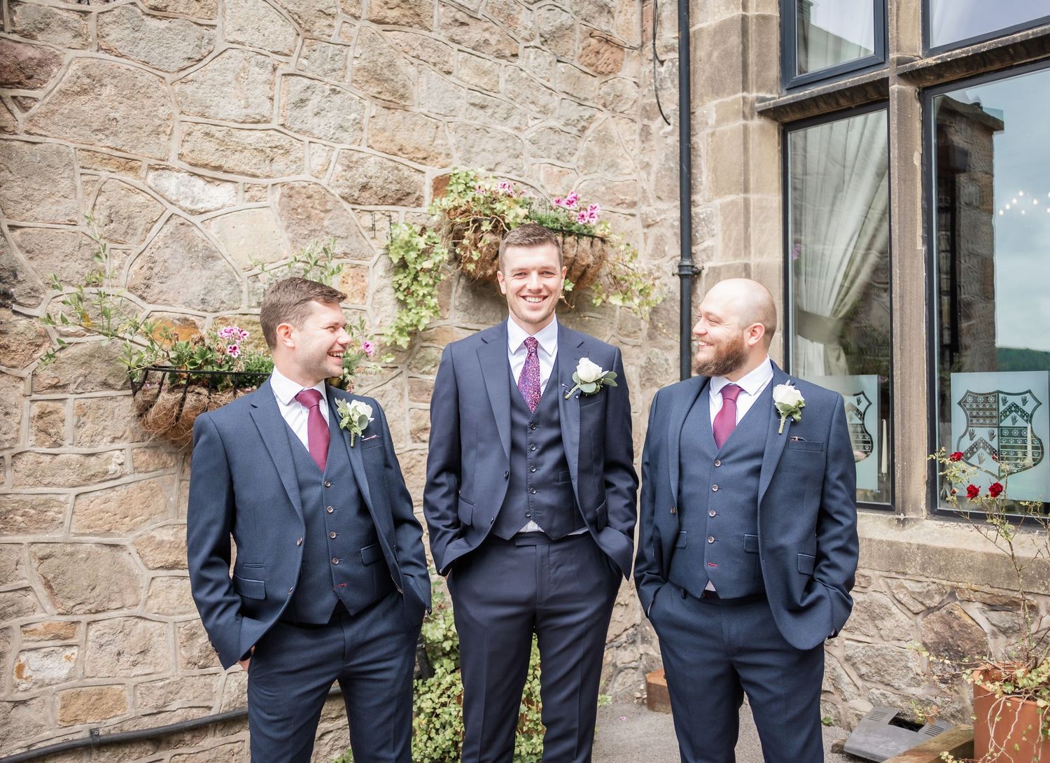 Three groomsmen in navy suits posing against stone wall exterior.