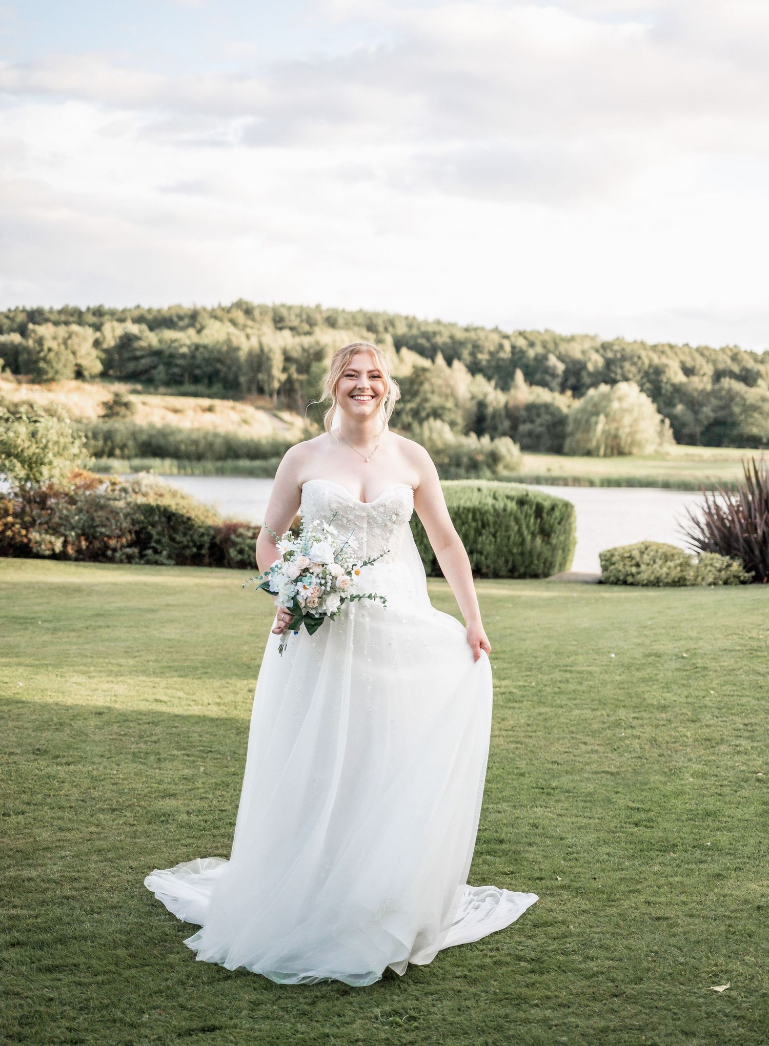Bride in a flowing white lace gown poses with bouquet on manicured lawn with lake view in background.
