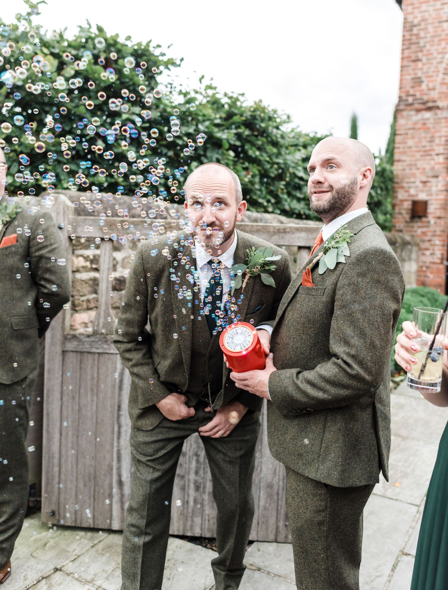 Two men in tweed suits enjoy bubbles during outdoor wedding festivities.