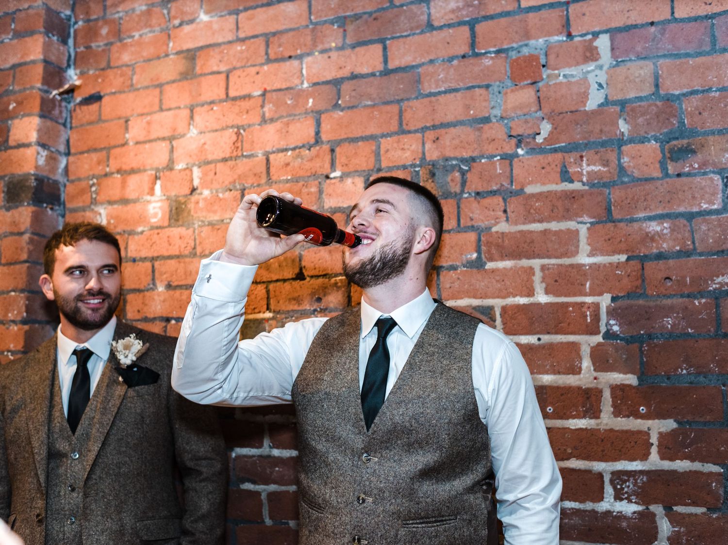 Two men in formal attire stand against brick wall while one drinks from bottle.