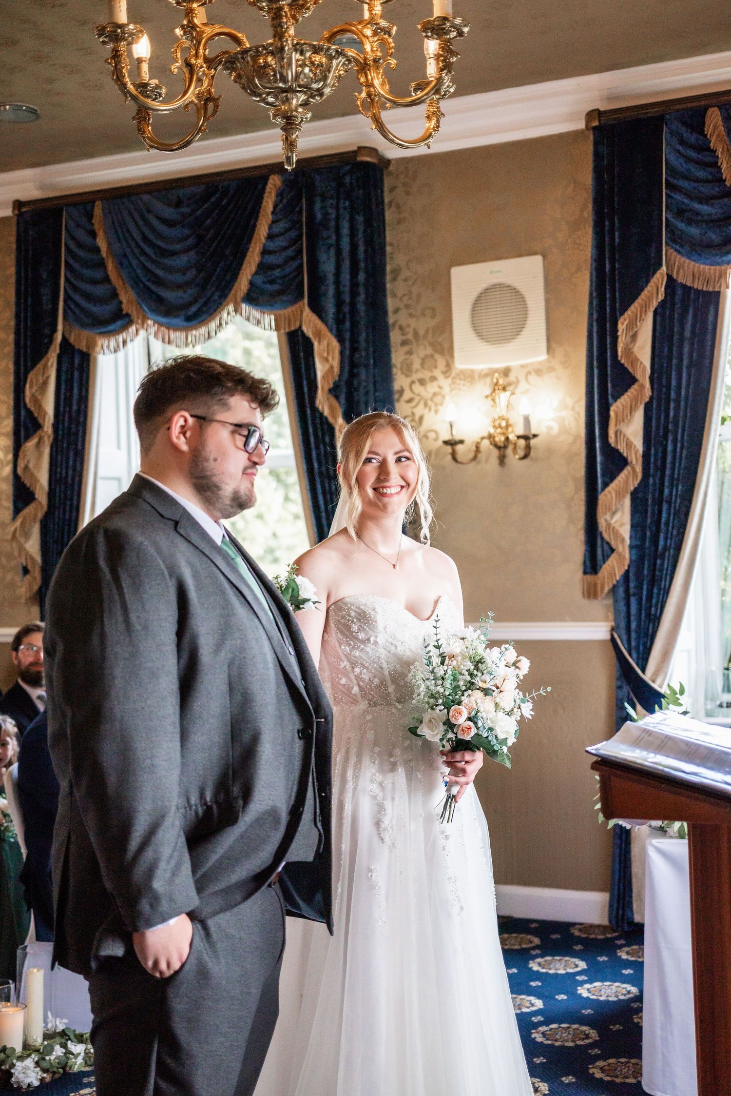Wedding couple standing in elegant room with navy curtains and chandelier overhead.