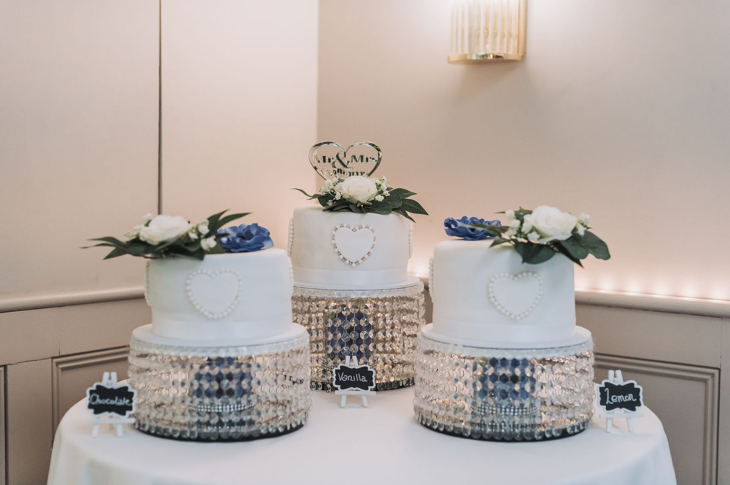 Three elegant white wedding cakes decorated with crystal details and greenery on display.
