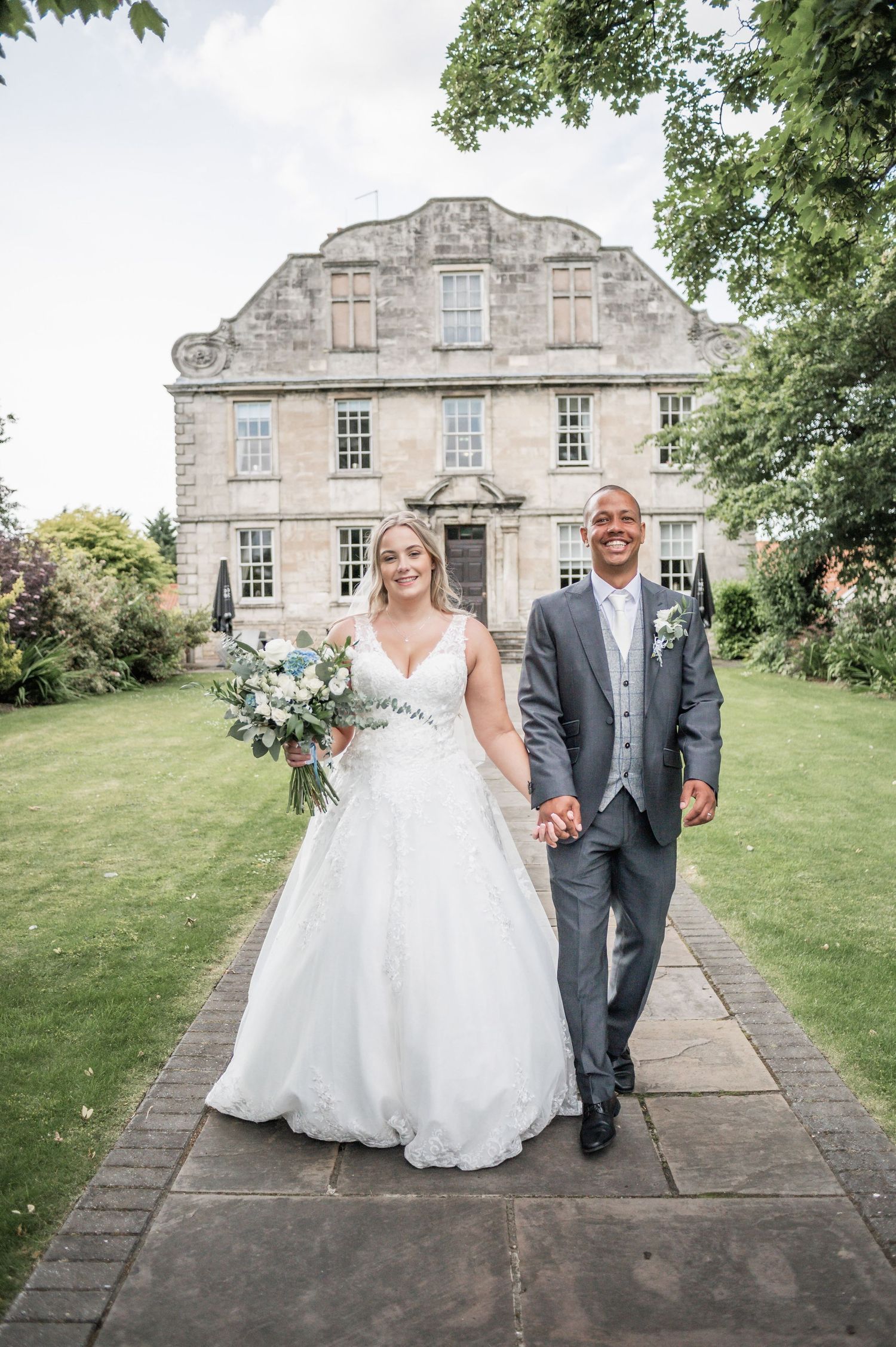 Newlyweds walk along garden path in front of elegant Georgian mansion.