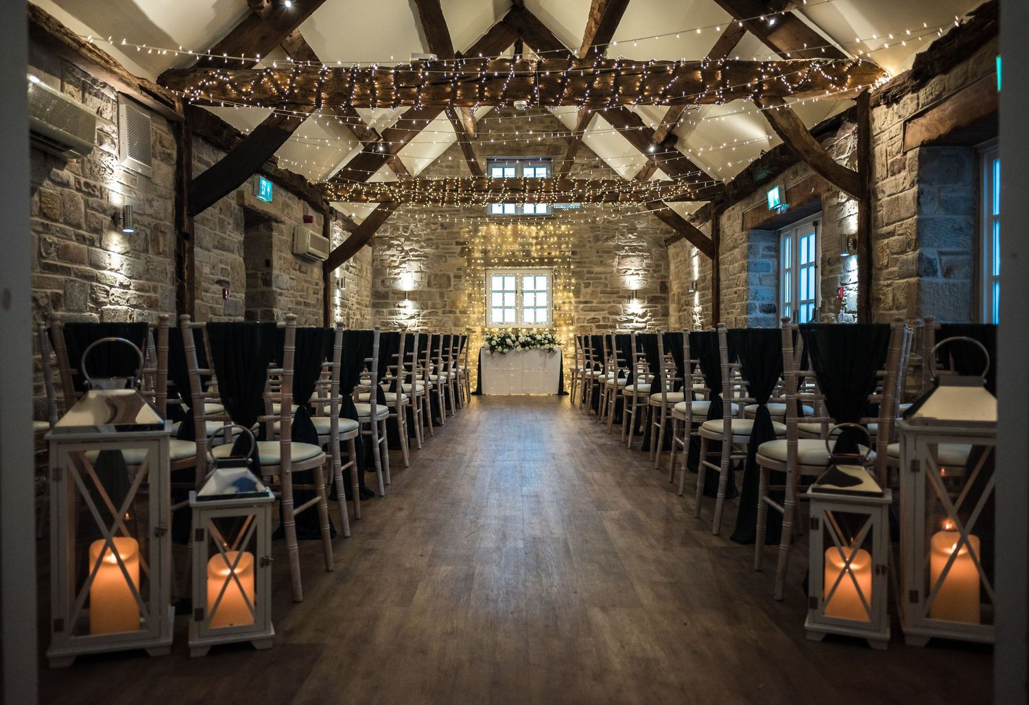 Rustic stone barn venue with wooden beams and lantern-lined aisle set up for a wedding ceremony.