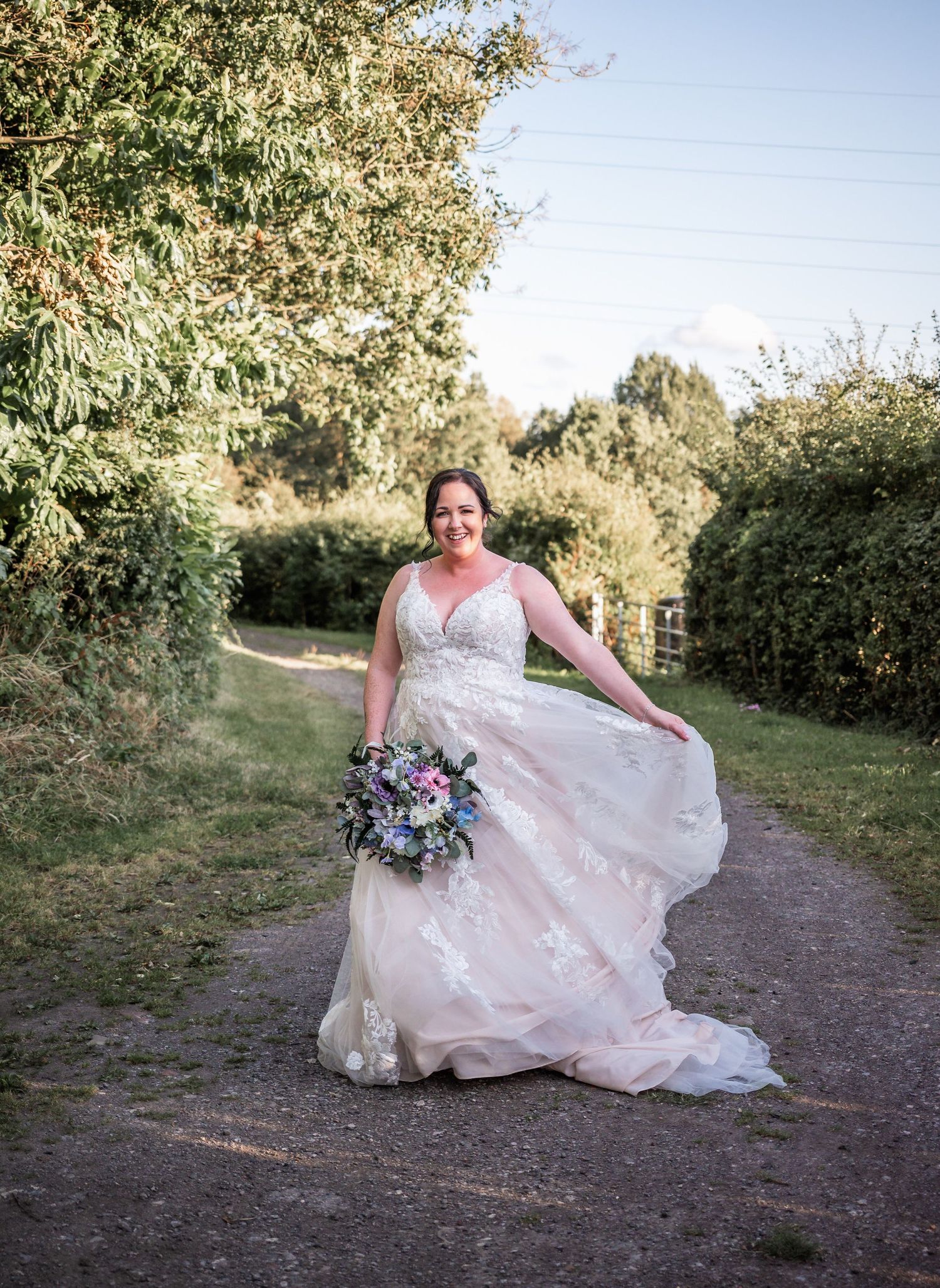 A bride in a lace wedding dress twirls on a garden path while holding a purple and blue bouquet.