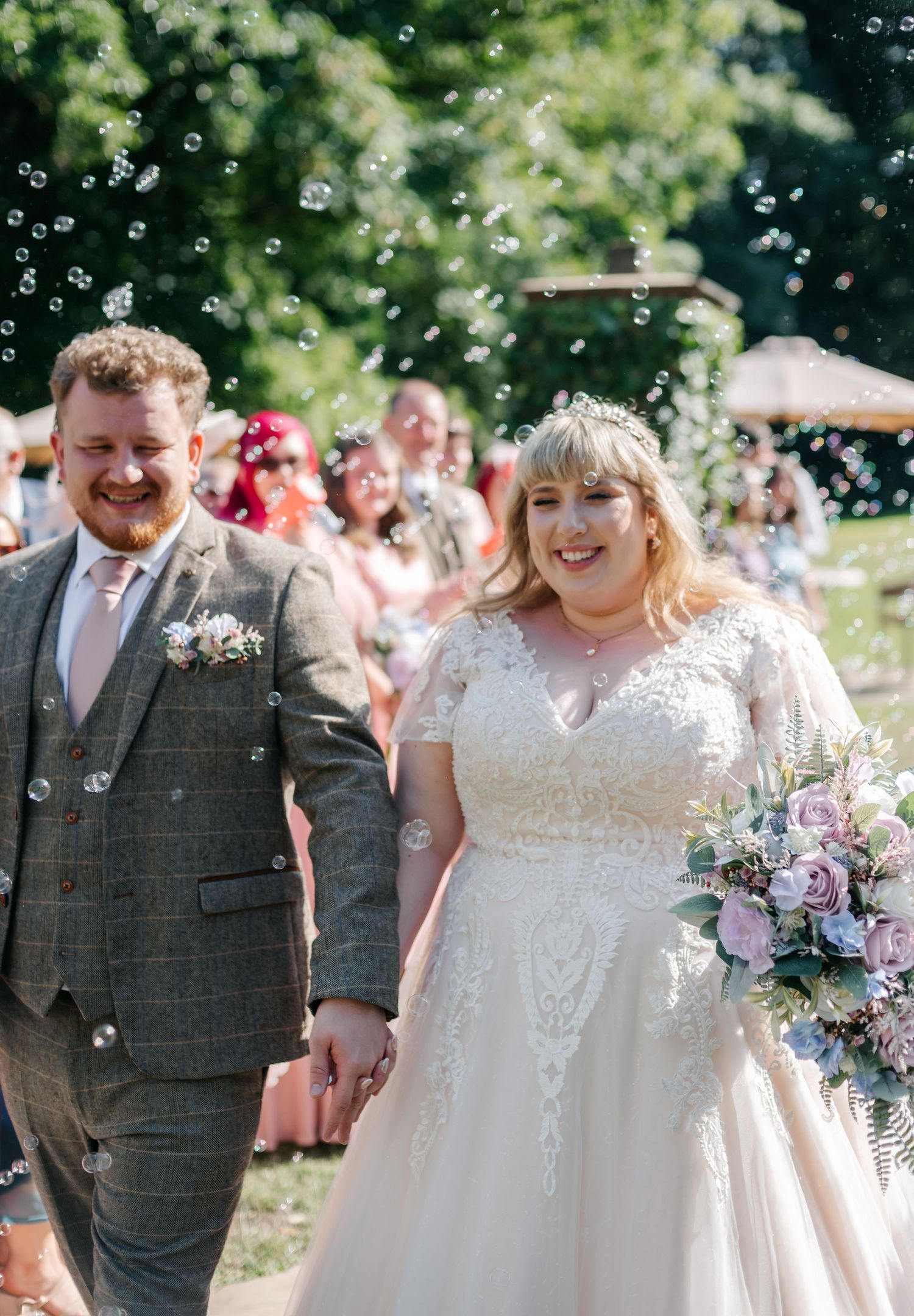 Newlyweds walking through bubbles after outdoor wedding ceremony, bride in lace gown holding pink bouquet.