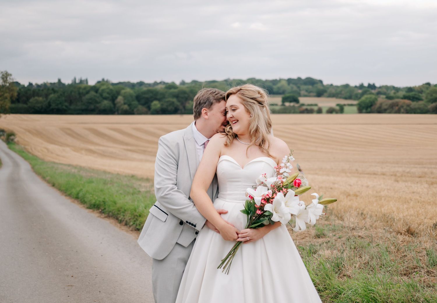 Newlyweds share romantic moment in countryside field with rolling hills backdrop.