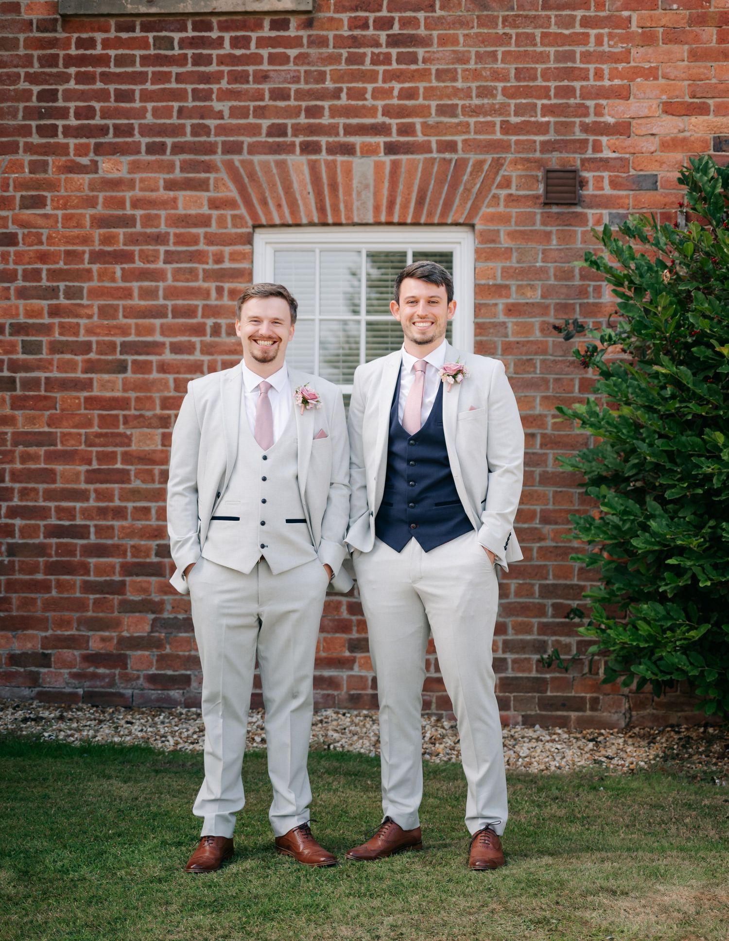 Groomsmen in light gray suits and navy vests pose casually against a red brick wall outdoors.