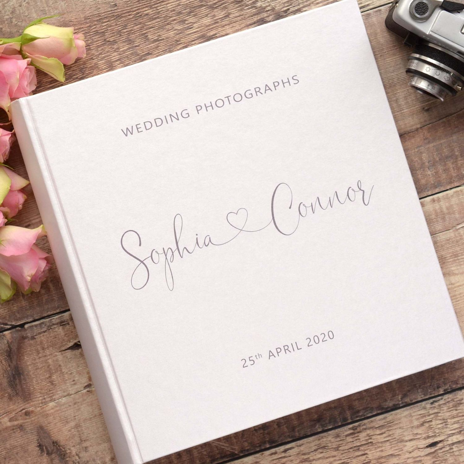 Elegant wedding photography album with white covers and calligraphy text displayed on rustic wooden surface with pink roses.